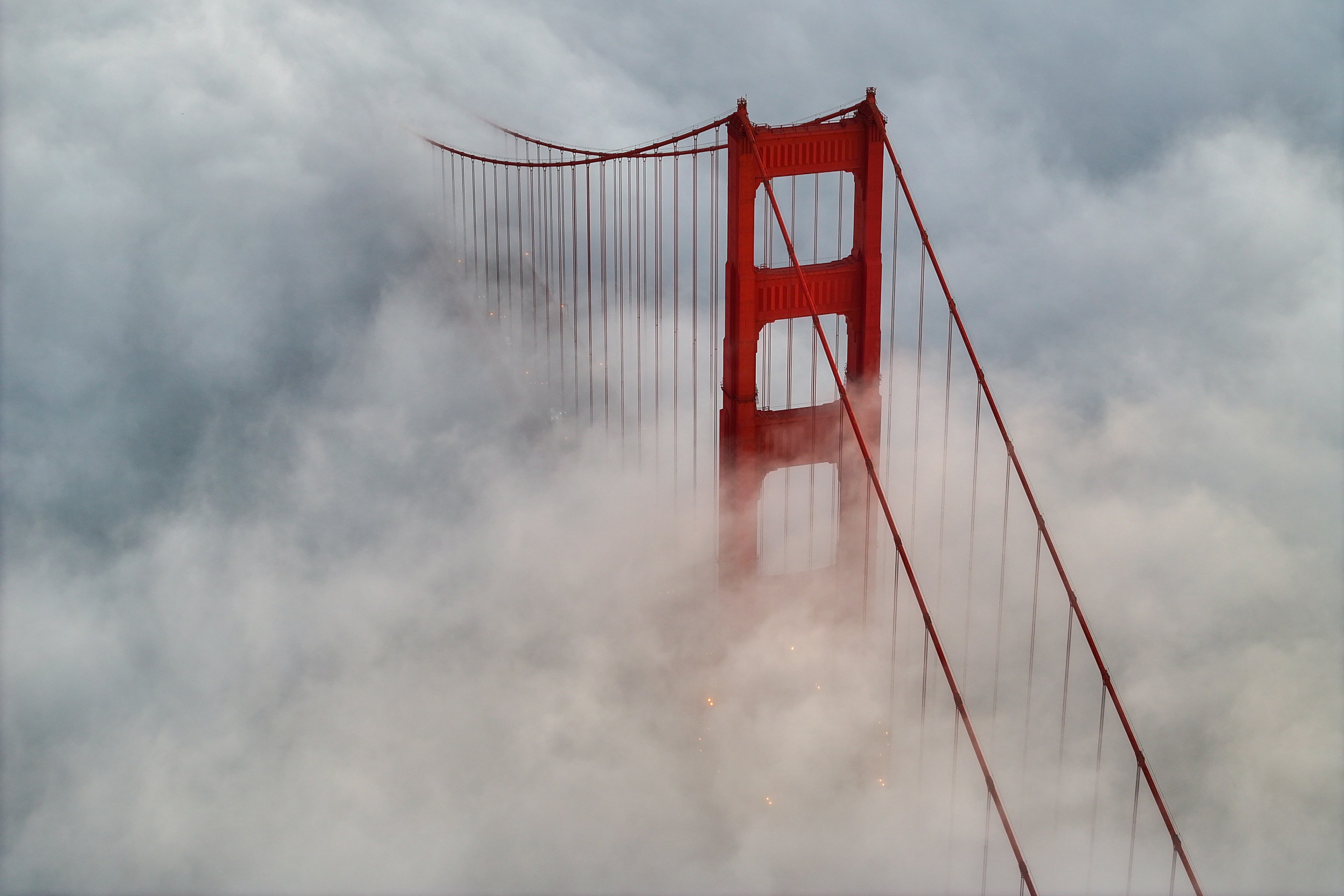 A tall, red suspension bridge partially shrouded in thick clouds, with its vertical towers and cables peeking through, creating an ethereal, misty scene.