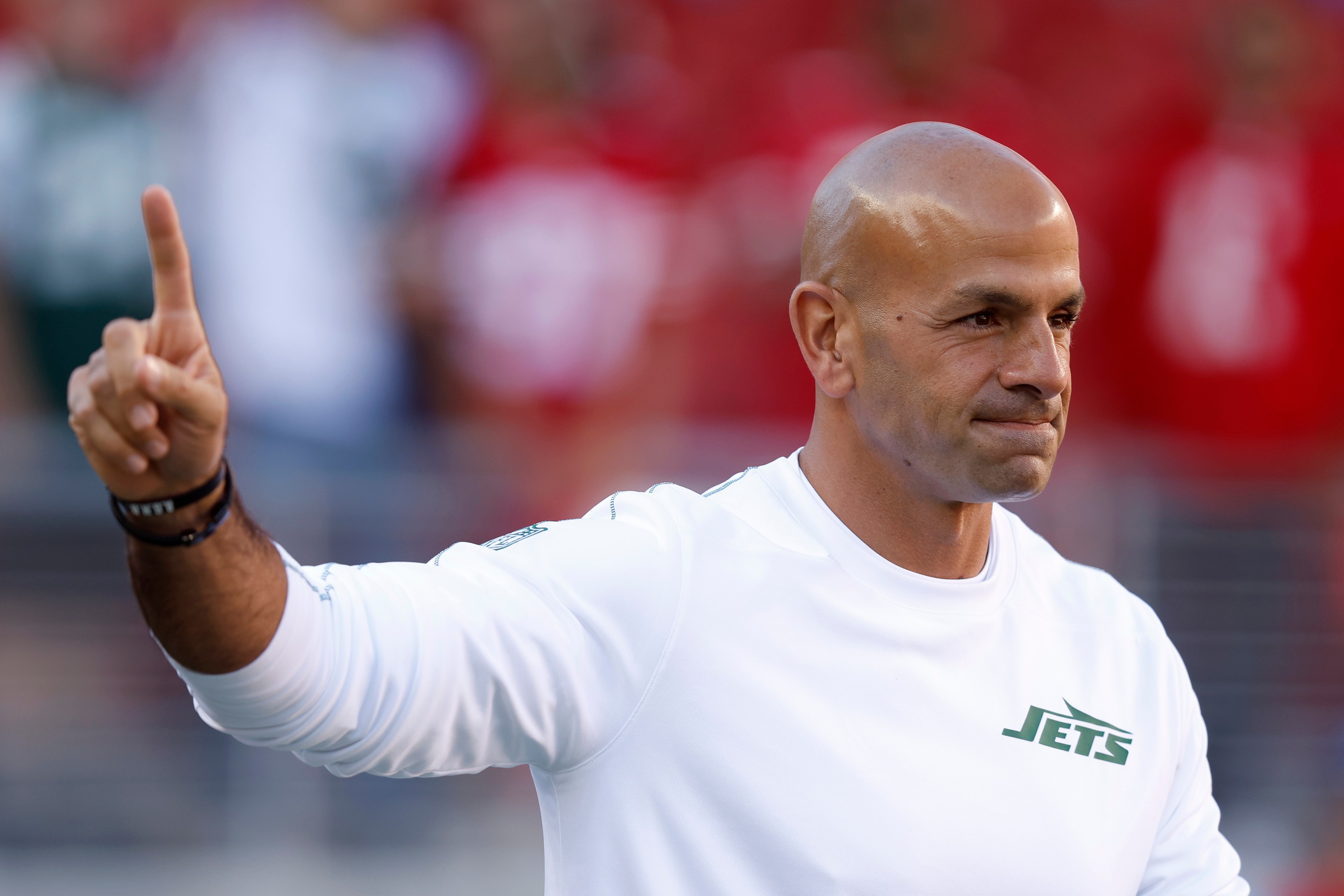 A man in a white Jets shirt raises his right hand with one finger up, while standing with a focused expression against a blurred red and white background.