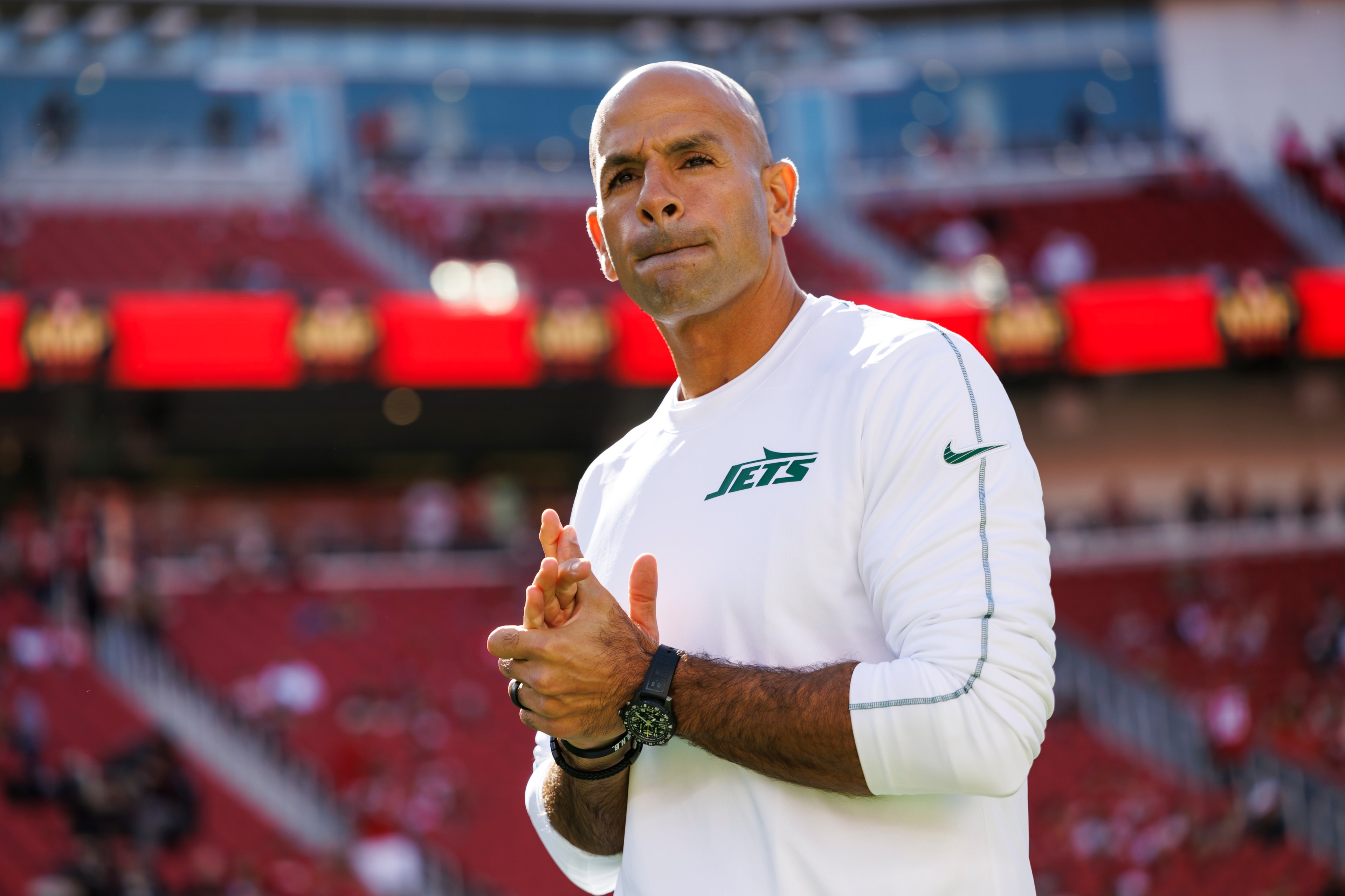 A person stands in a stadium wearing a white Jets shirt, with their hands clasped. The background shows blurred red stadium seats.