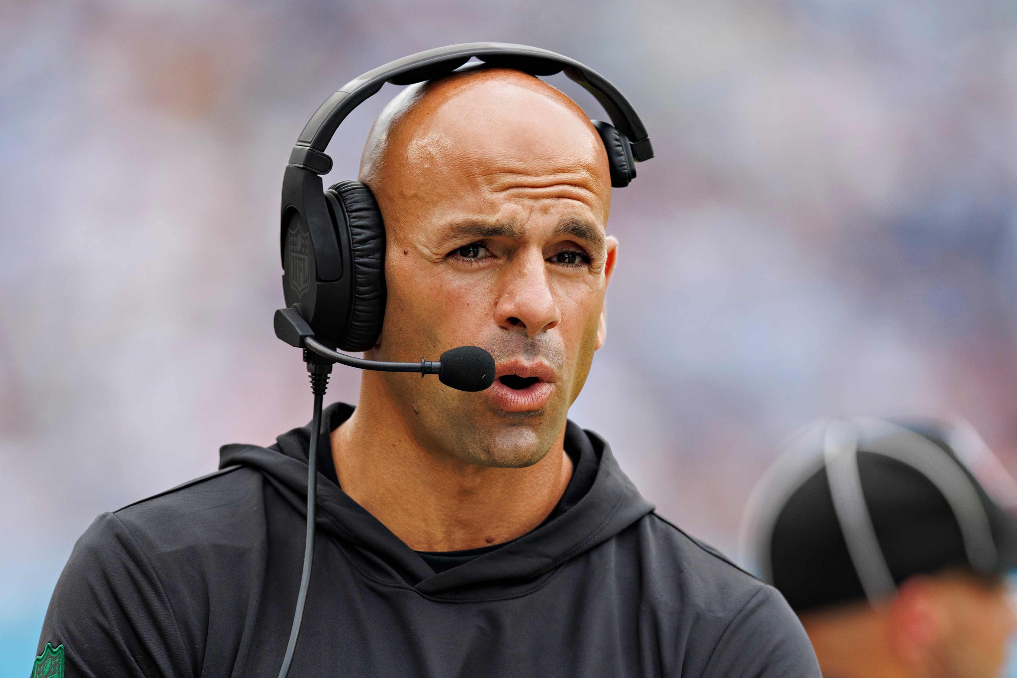 A person wearing a headset and a black jacket appears to be speaking, likely in a coaching or commentary role, with a blurred crowd in the background.