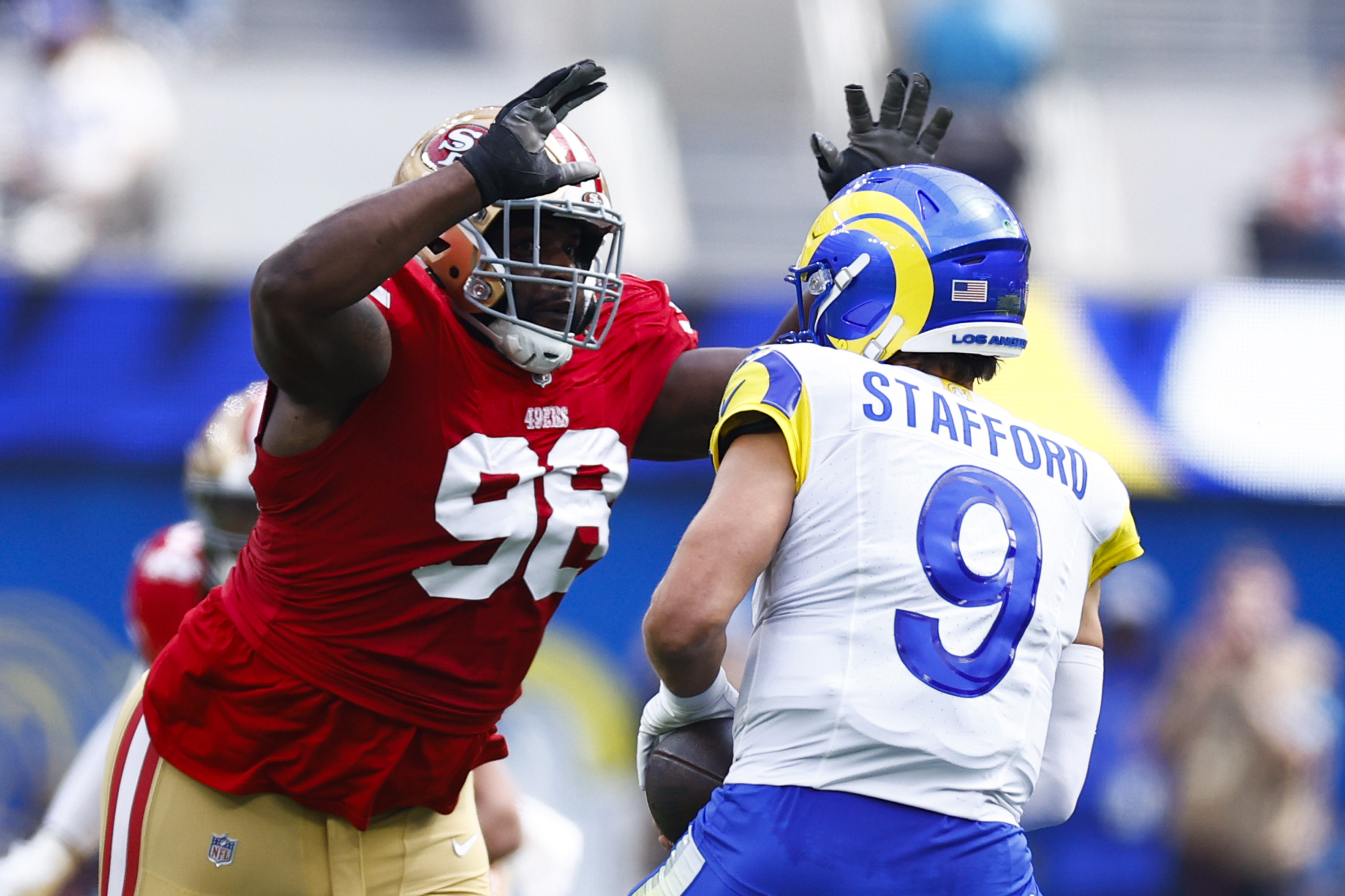 A football player in a red jersey raises his arms, facing another in a white and blue jersey holding a ball, suggesting an action-packed game moment.