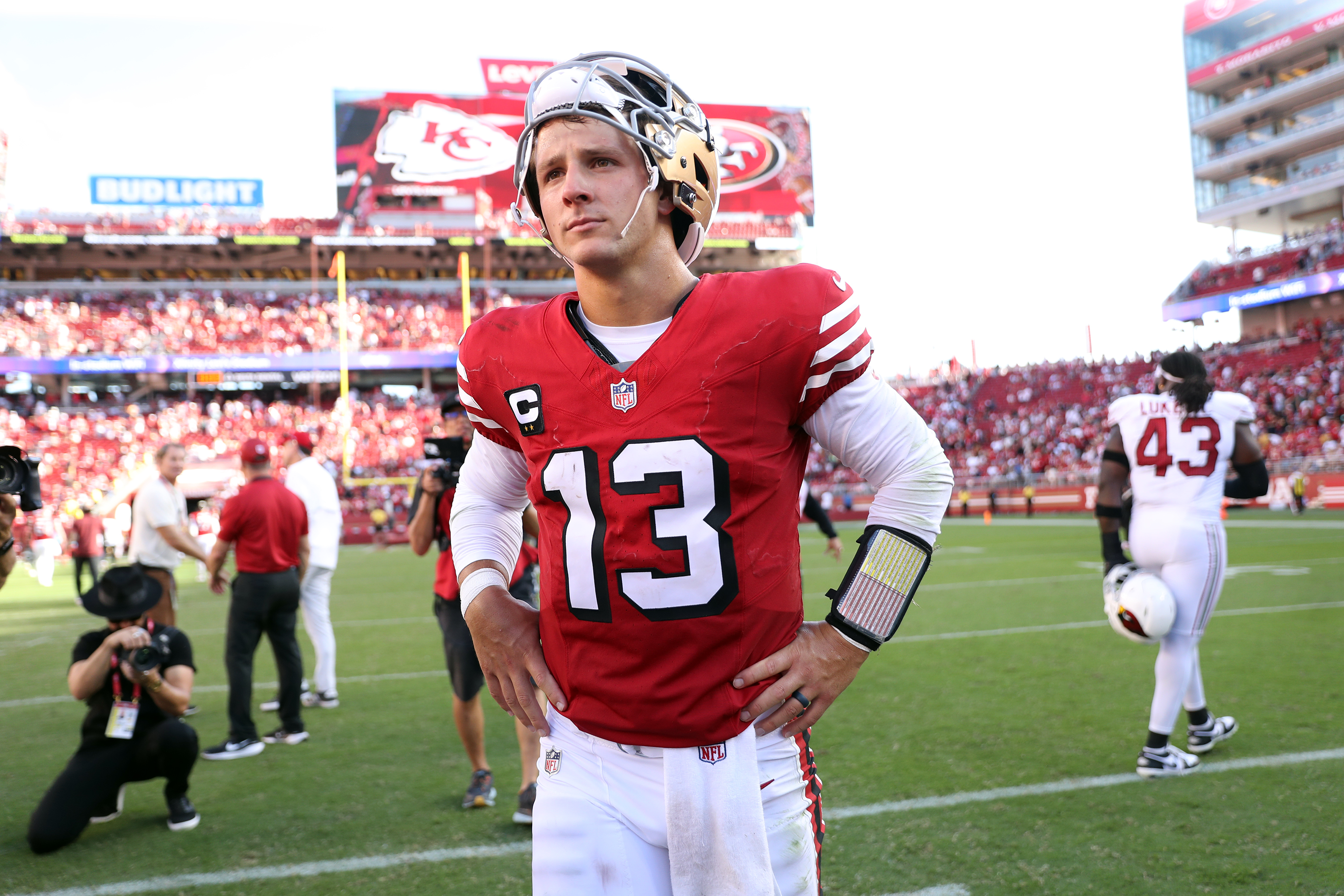 A football player in a red jersey with the number 13 stands on the field, hands on hips, with a helmet on and a crowded stadium in the background.