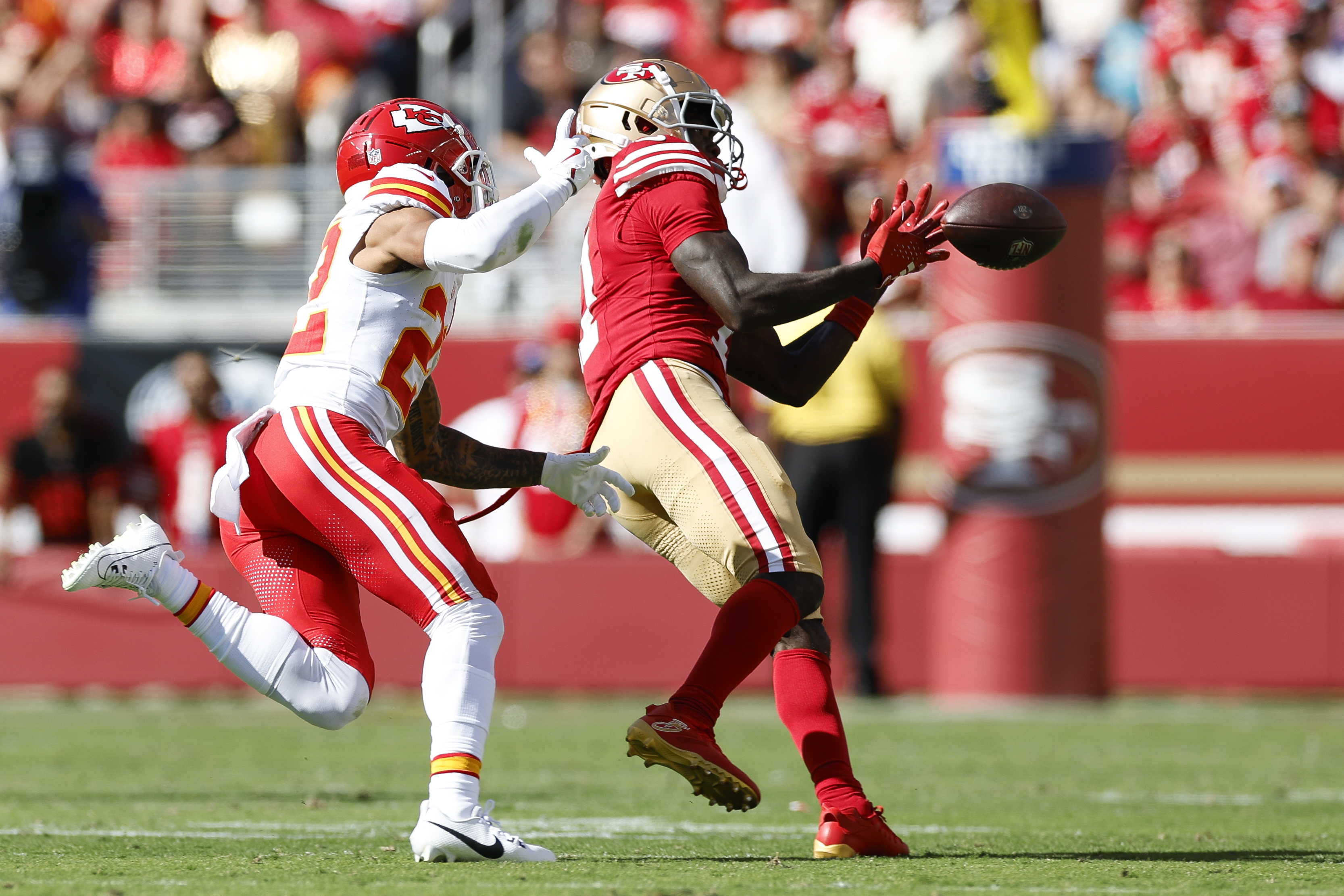 Two football players are in action on a field. One in a red and gold uniform is reaching to catch the ball, while the other in a red and white uniform defends.