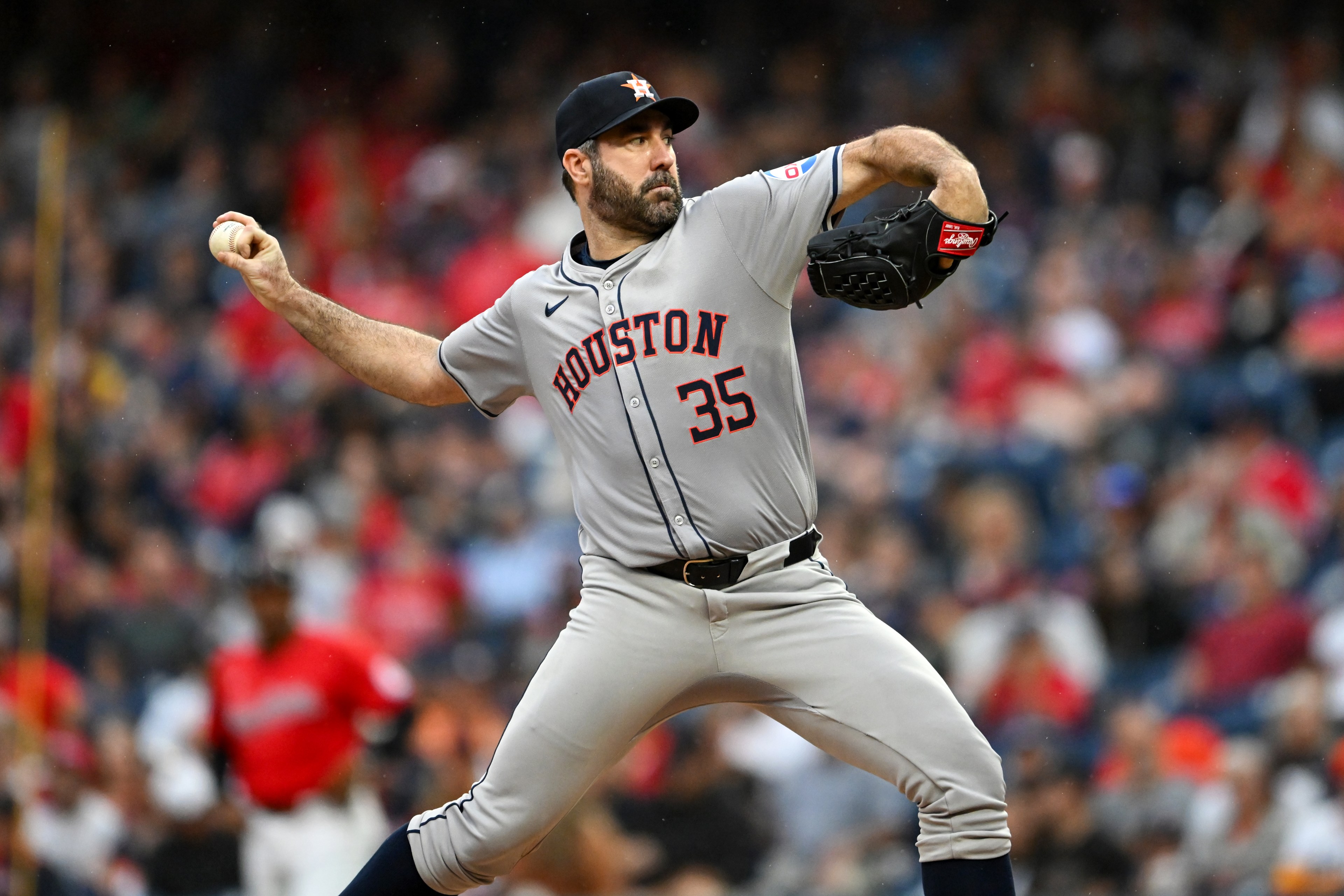 A baseball player in a gray "Houston" uniform pitches on a field, captured mid-throw. The crowd in the background is blurred.