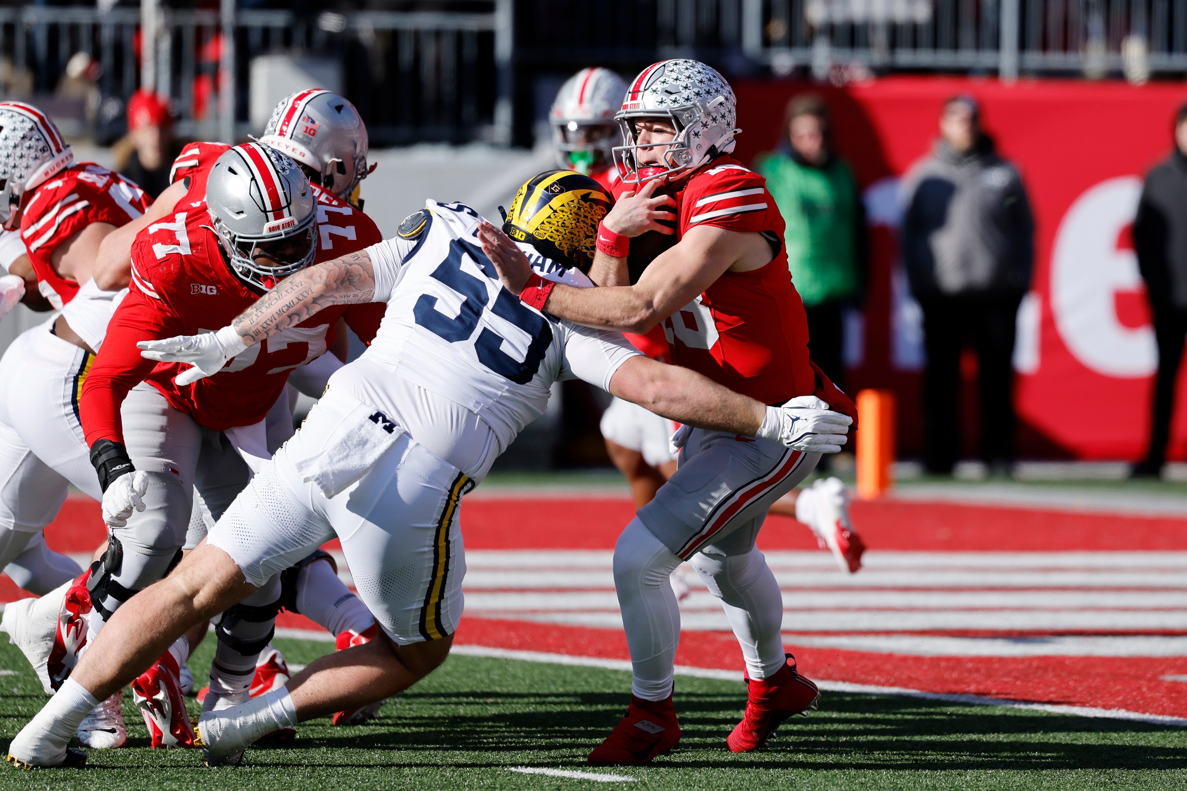 Two football players collide near an end zone. One wears a red and gray uniform, holding the ball; the other, in white and yellow, attempts a tackle.