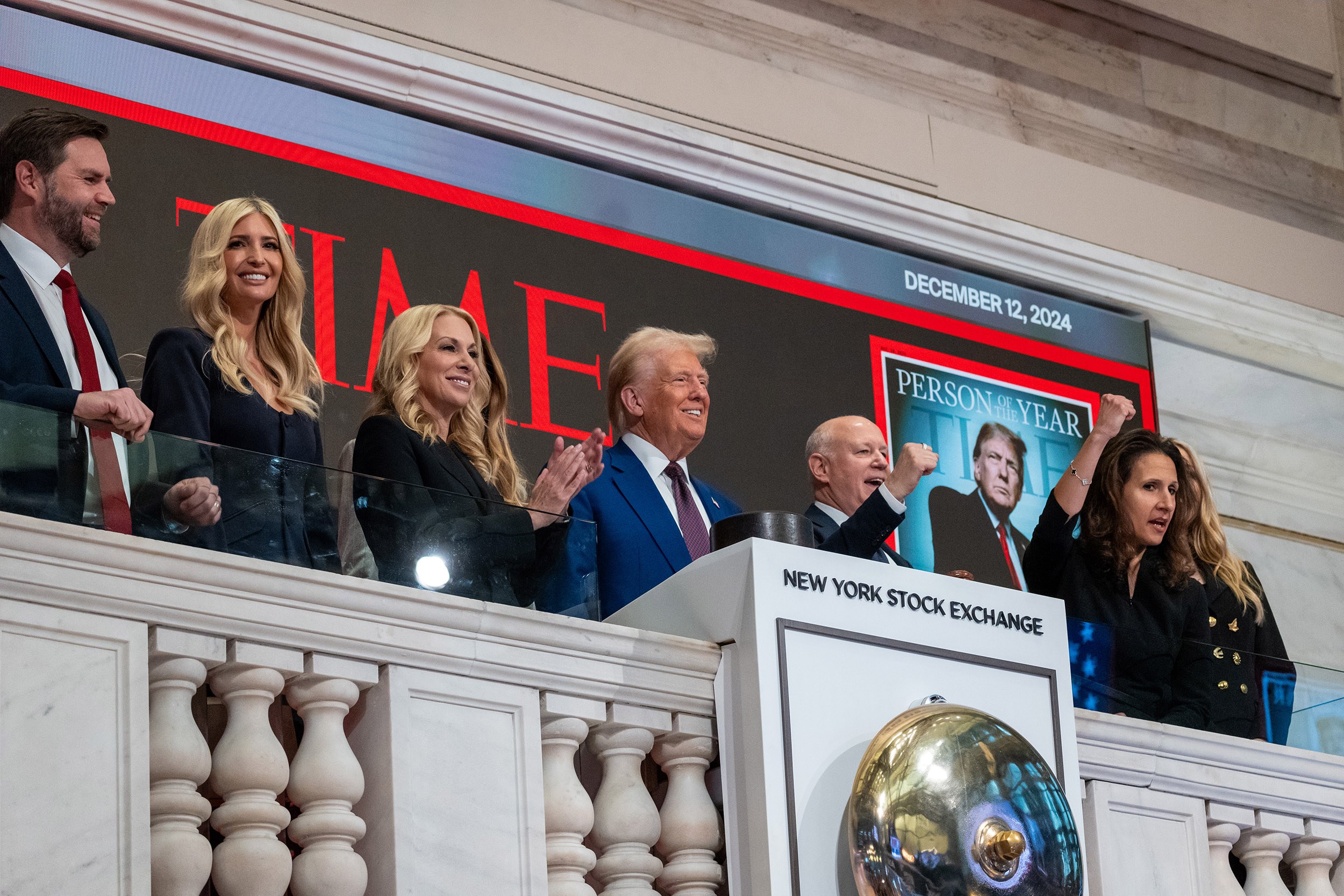 A group of people are standing on a balcony at the New York Stock Exchange. They're smiling and applauding, with a large &quot;Time Person of the Year&quot; display behind them.