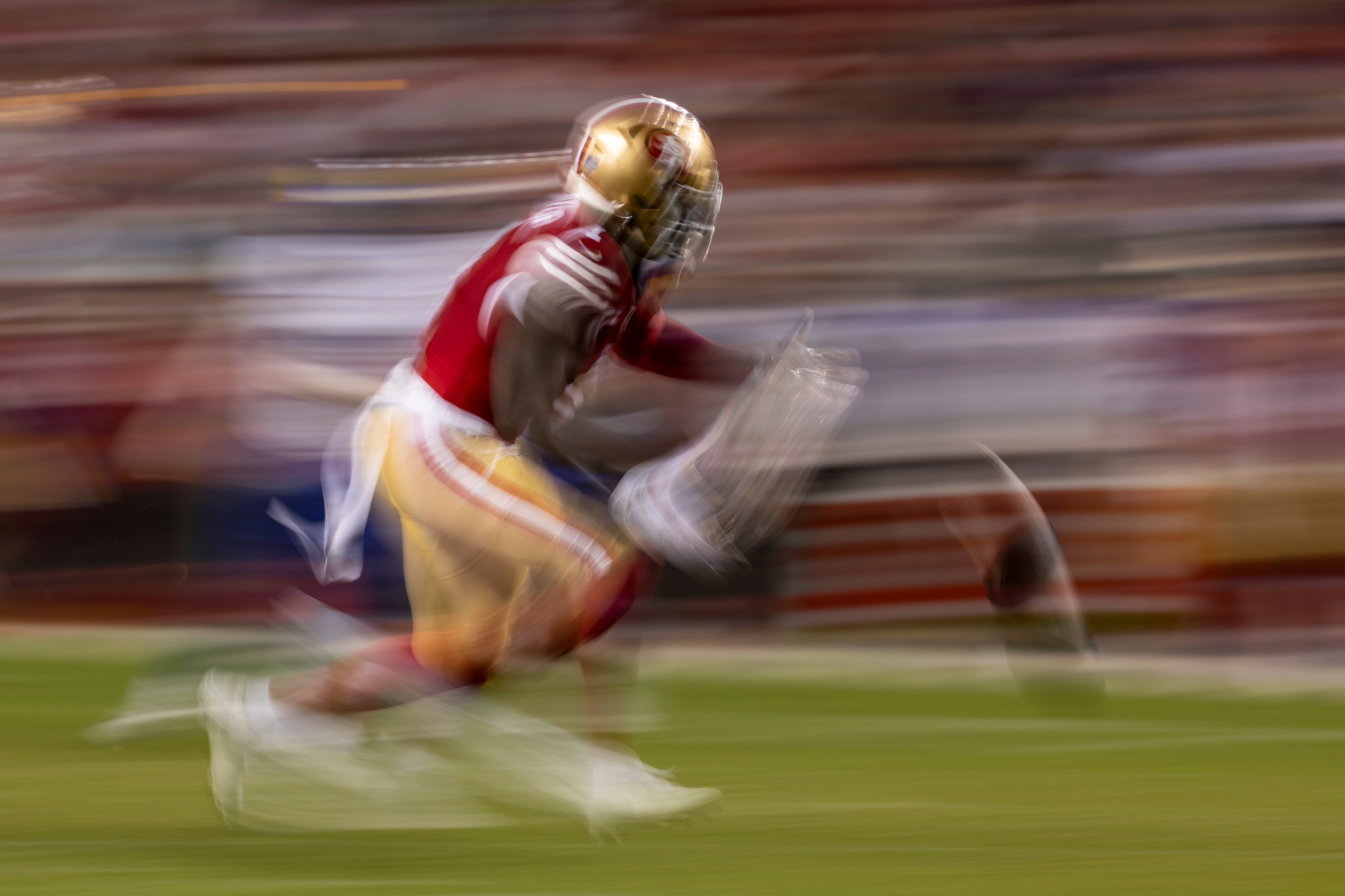 A football player in red and gold, blurred in motion, is running quickly on a field. The background is streaked, indicating speed.