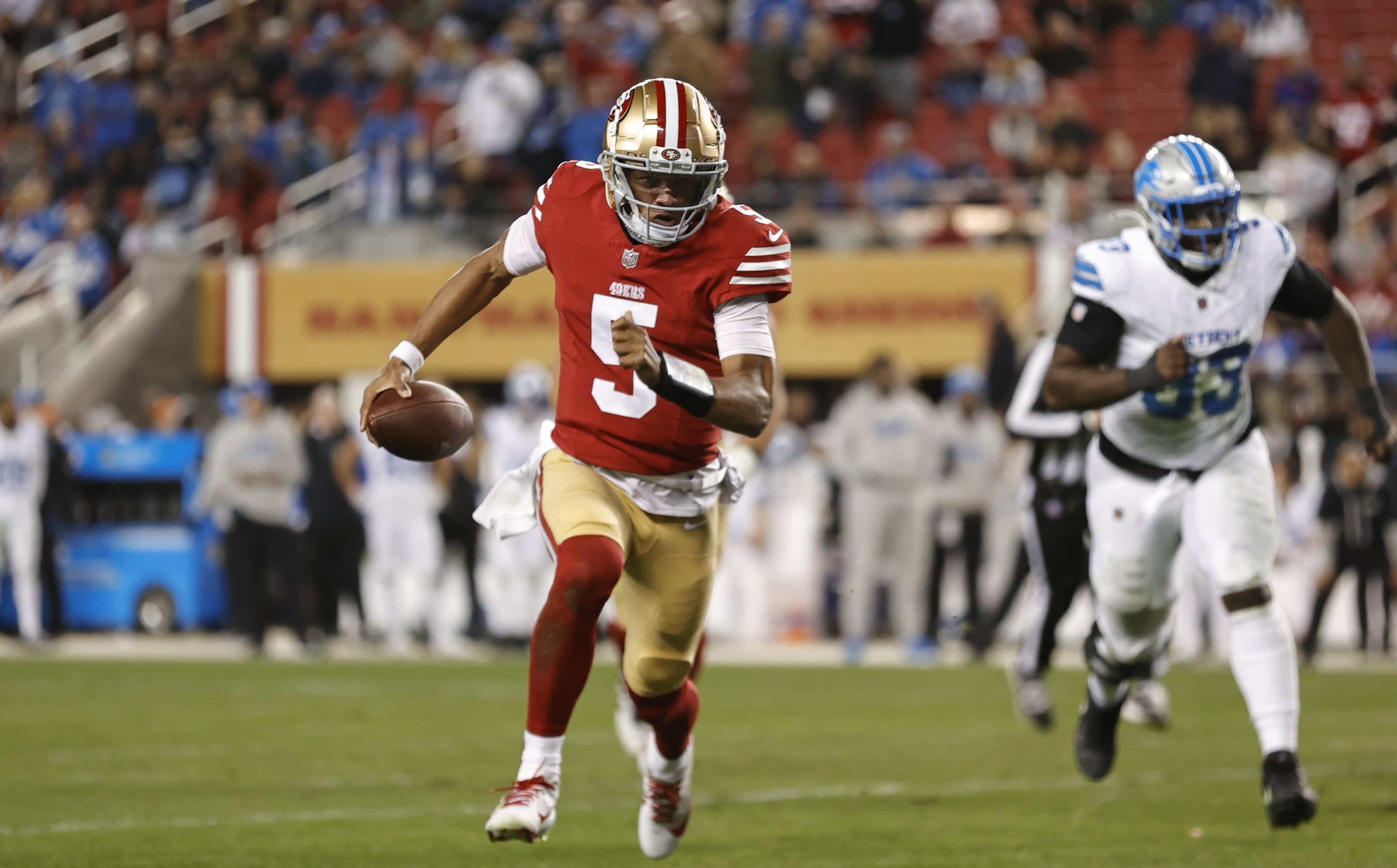 A football player in a red and gold uniform is running on the field with the ball. A player in a white and blue uniform is chasing him, with a crowd in the background.