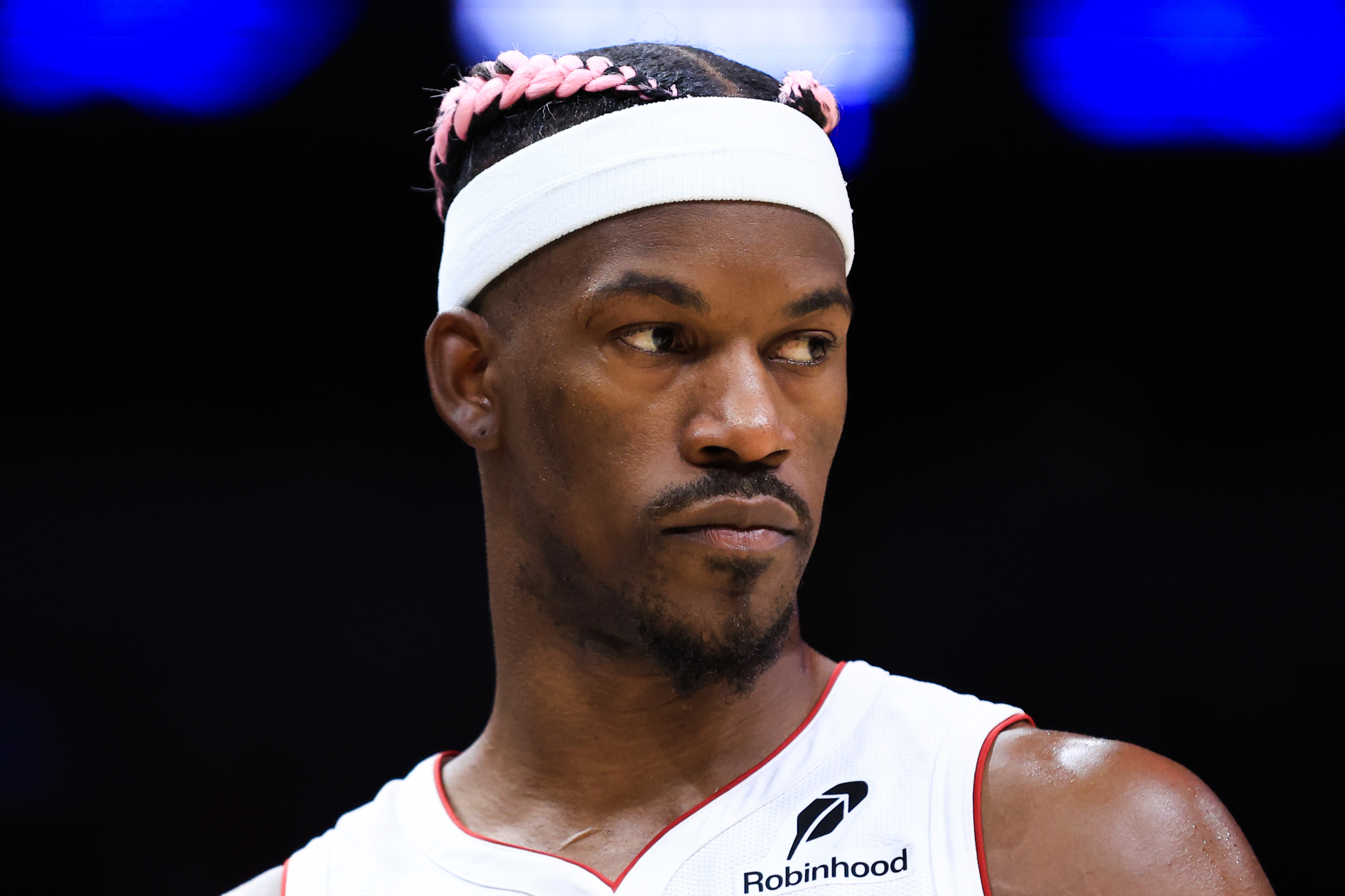 A basketball player wearing a white jersey and headband with pink hair twists looks to the side, displaying a focused expression against a dark background.