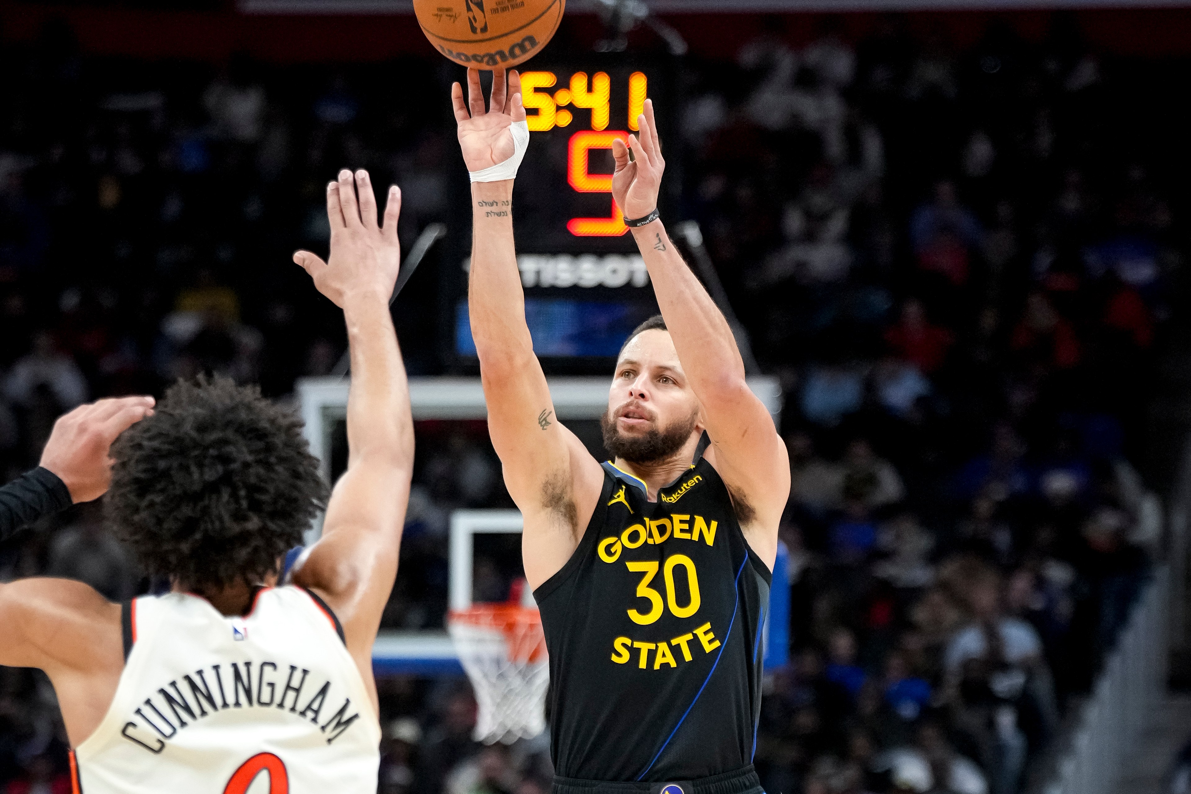 A basketball player in a black Golden State jersey shoots a ball mid-air while another player in a white jersey defends him. The crowd is visible in the background.