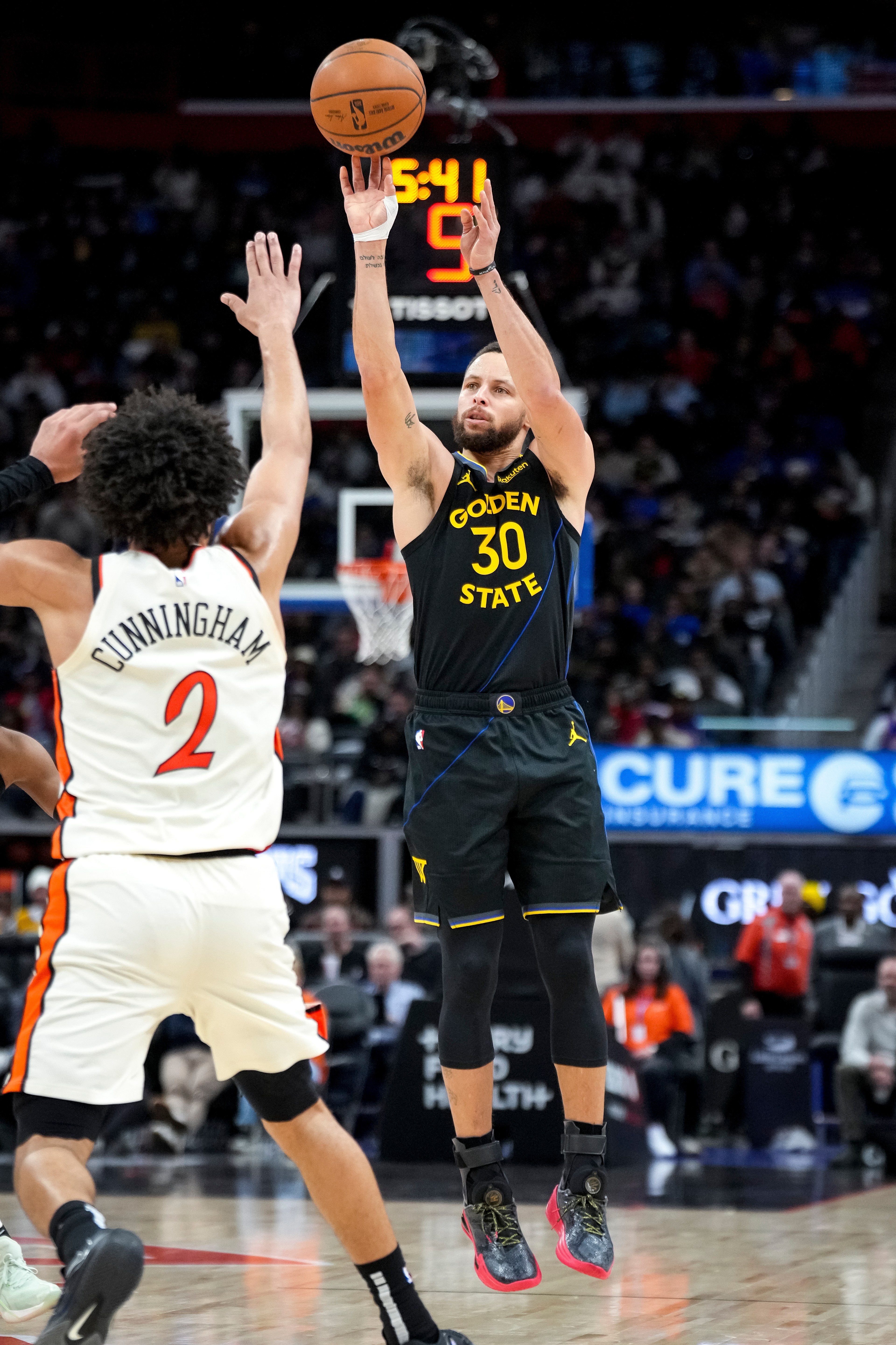 A basketball player in a black Golden State jersey shoots a ball mid-air while another player in a white jersey defends him. The crowd is visible in the background.