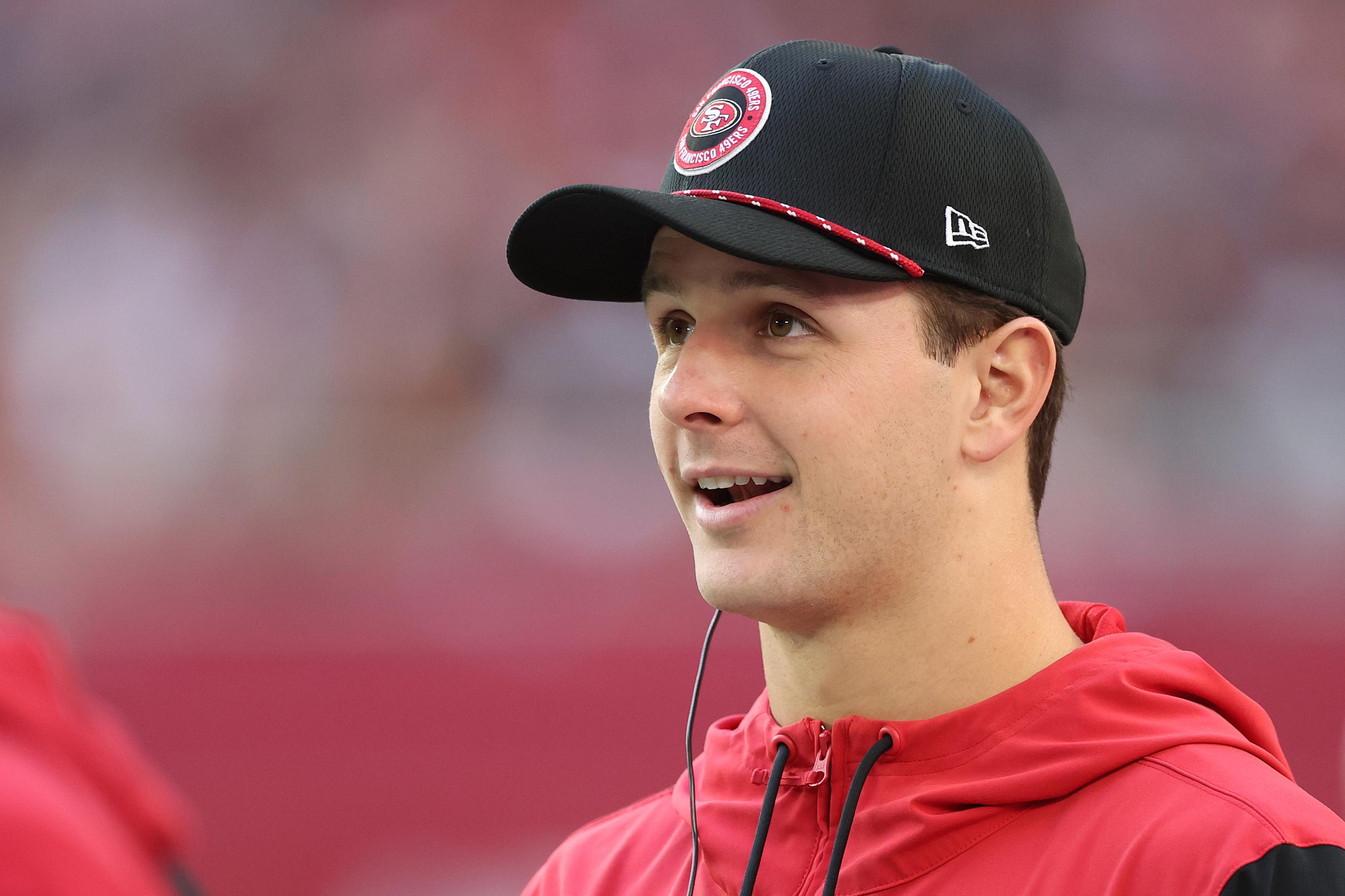 A person in a red jacket and black cap with a logo is looking up with a surprised expression. The background is blurred, suggesting an outdoor setting.