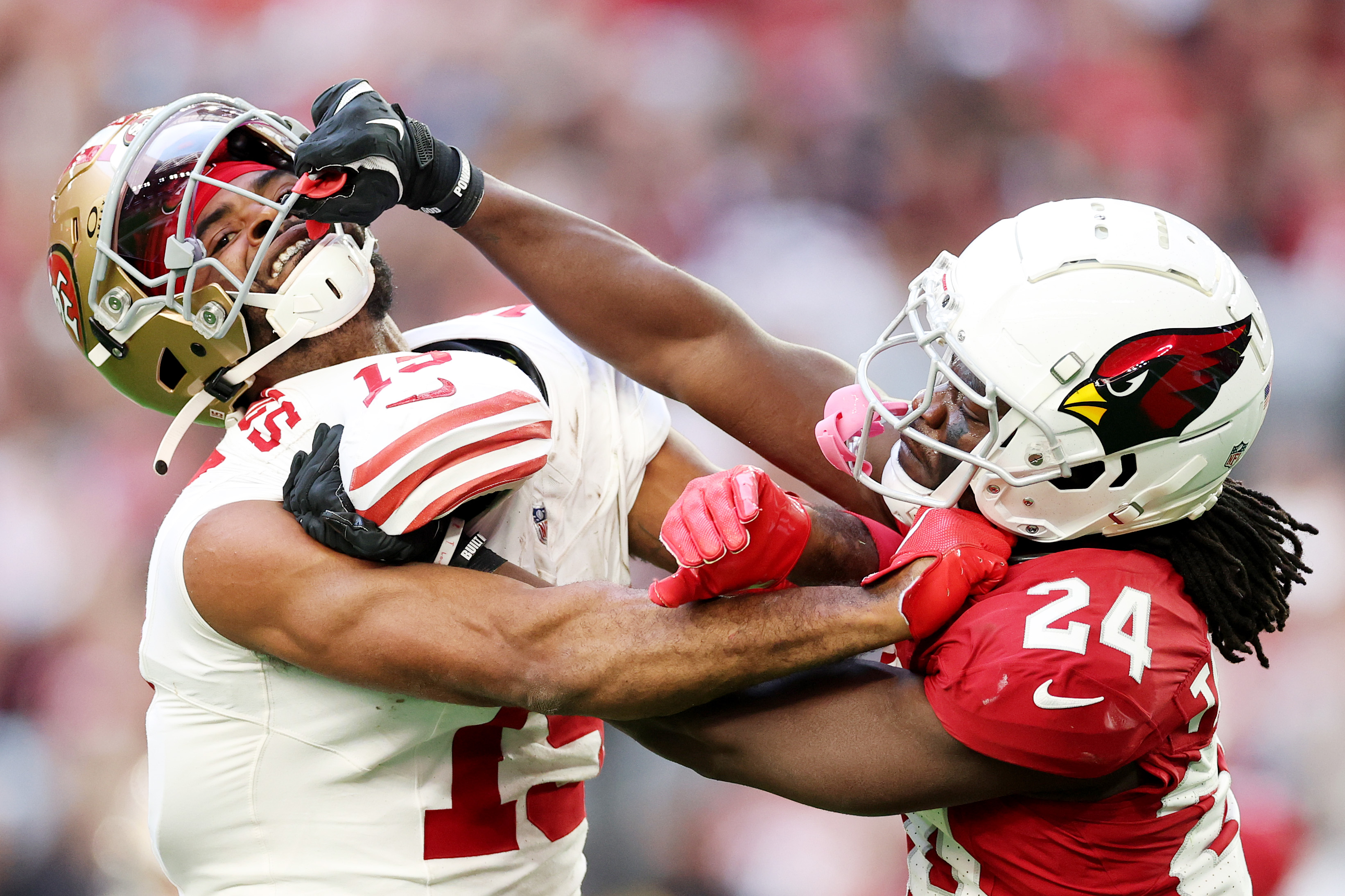 Two football players are engaged on the field. One player, in a white jersey, has his face mask grabbed by a player in a red jersey with a bird logo helmet.