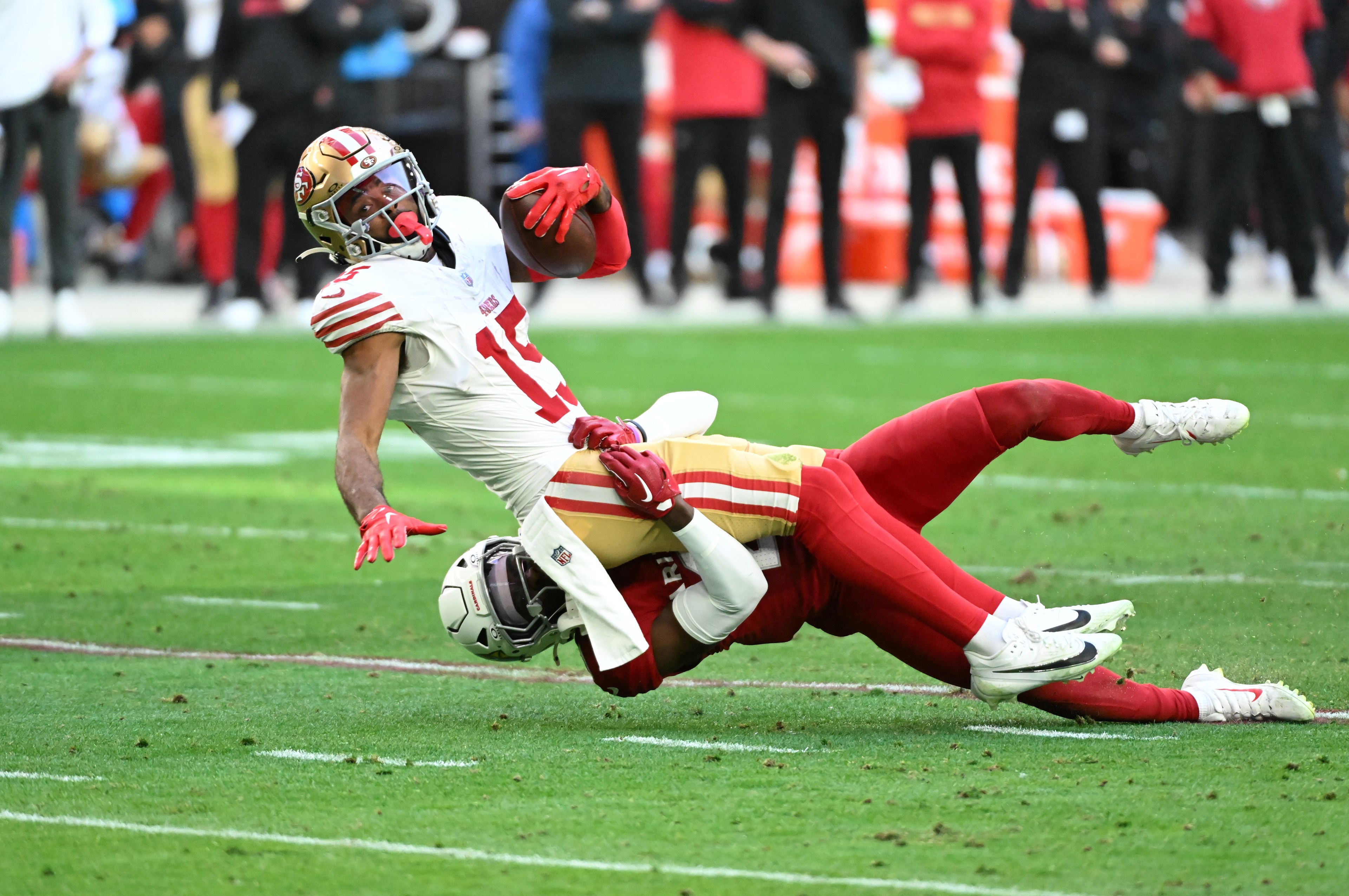 A football player in a white and red uniform is tackled to the ground by a player in red and white. The player holds the ball tightly during the tackle.