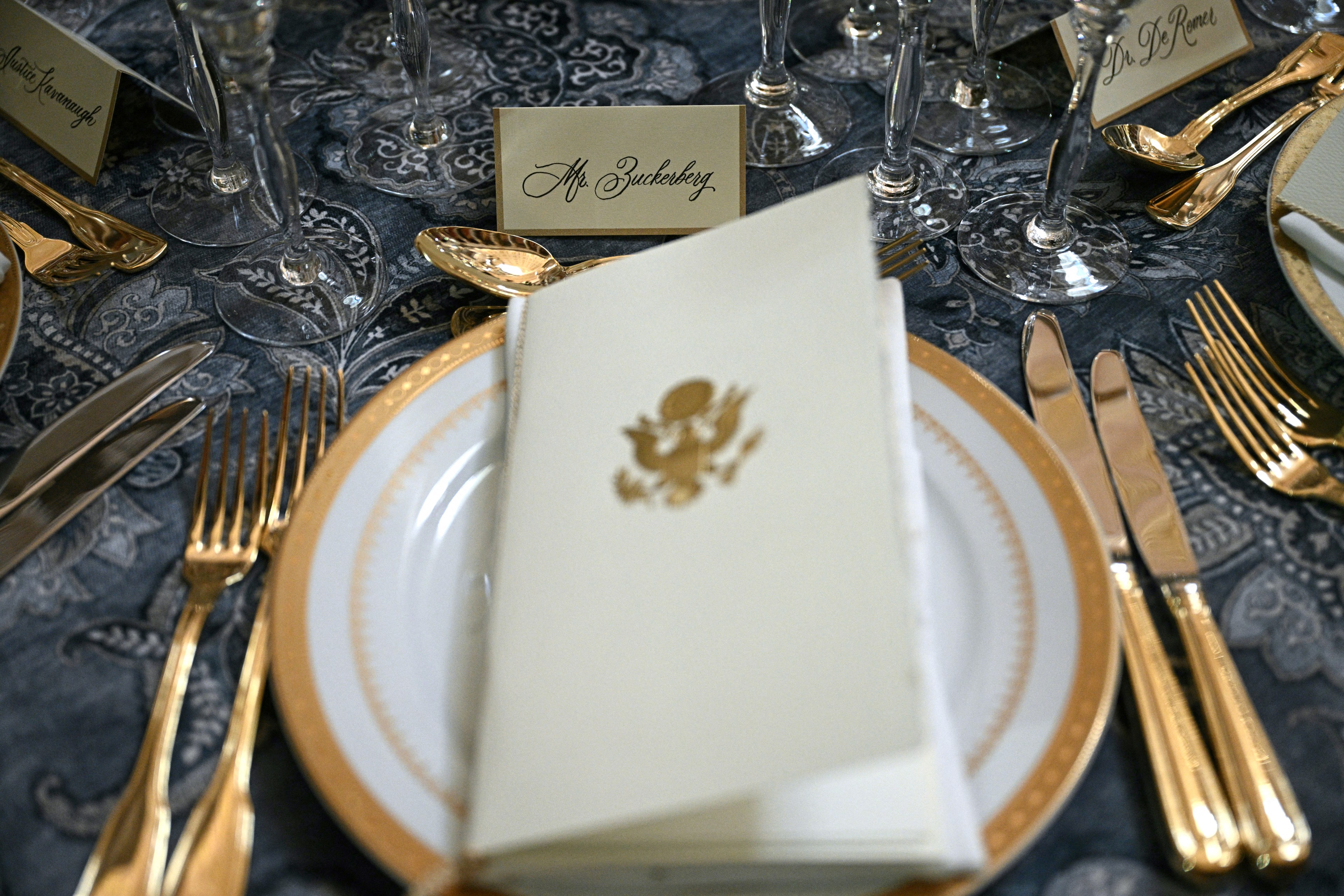 A formal dinner setup features gold-rimmed plates, elegant cutlery, and glassware on a patterned cloth. A folded napkin with a gold emblem and name cards are present.