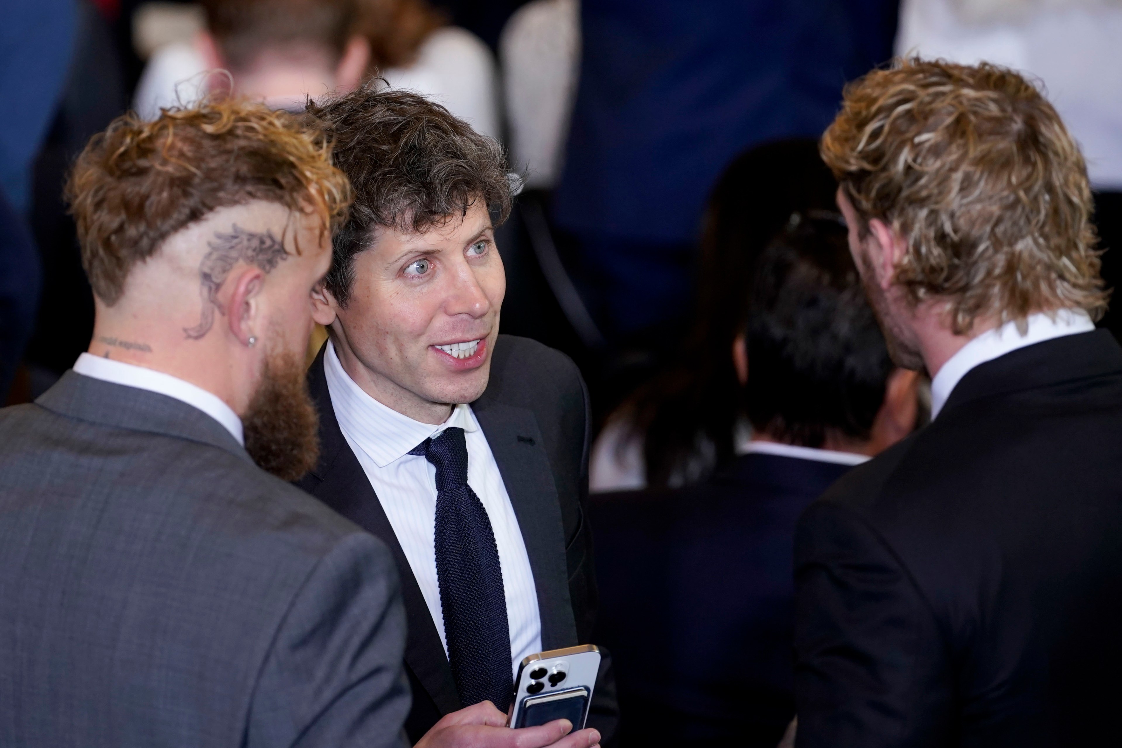 Three men in suits are engaged in conversation. One has a tattoo on his head, another holds a phone. They appear to be in a crowded social setting.