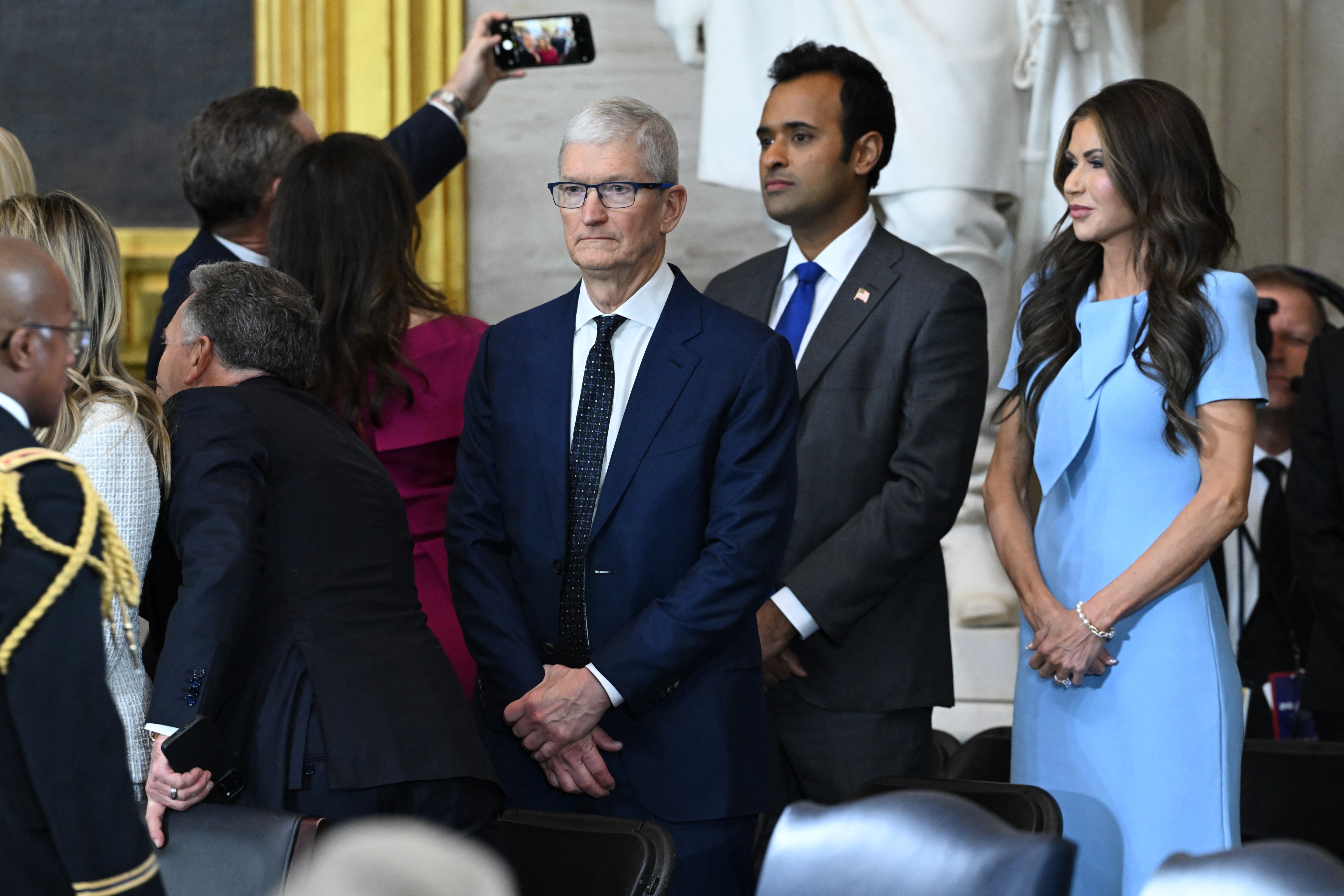 A group of people stand in a formal setting. One person takes a selfie, while others are dressed in business attire. A woman in a light blue dress is visible.