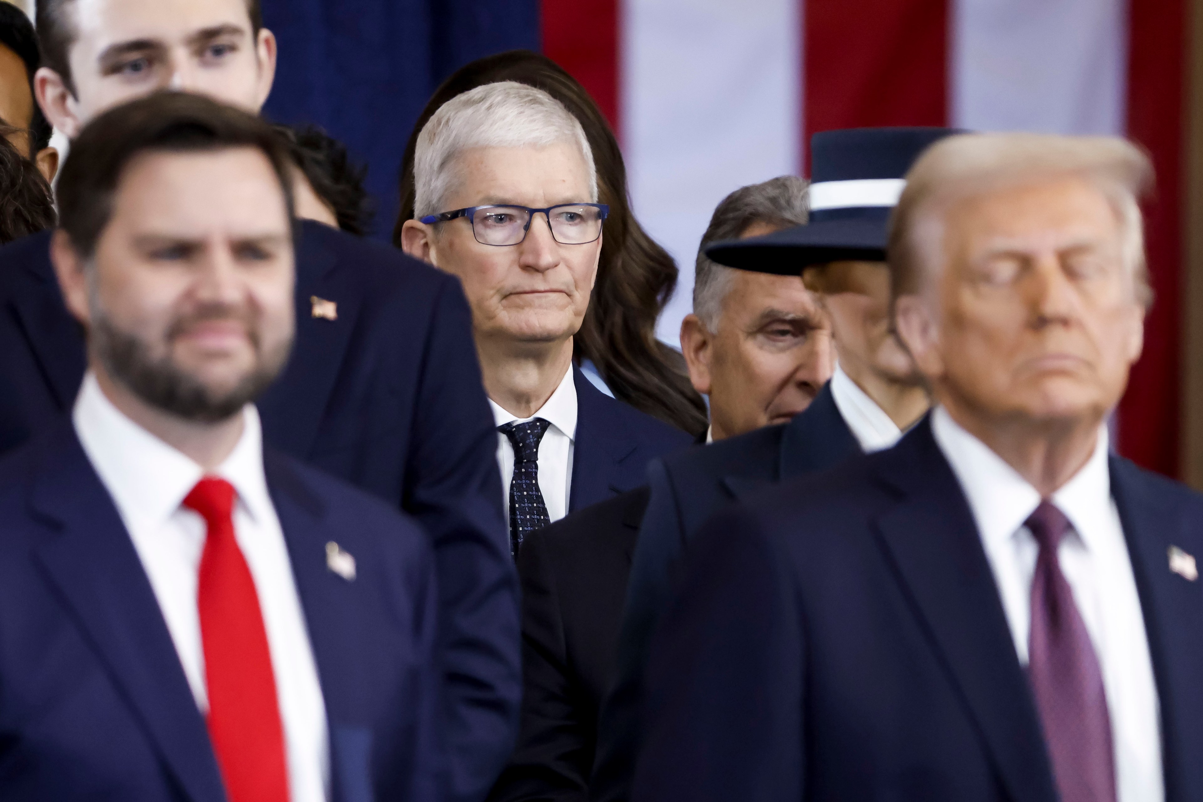 A group of men in suits, some with serious expressions, stand closely together. One wears glasses. A blurred striped background is visible.