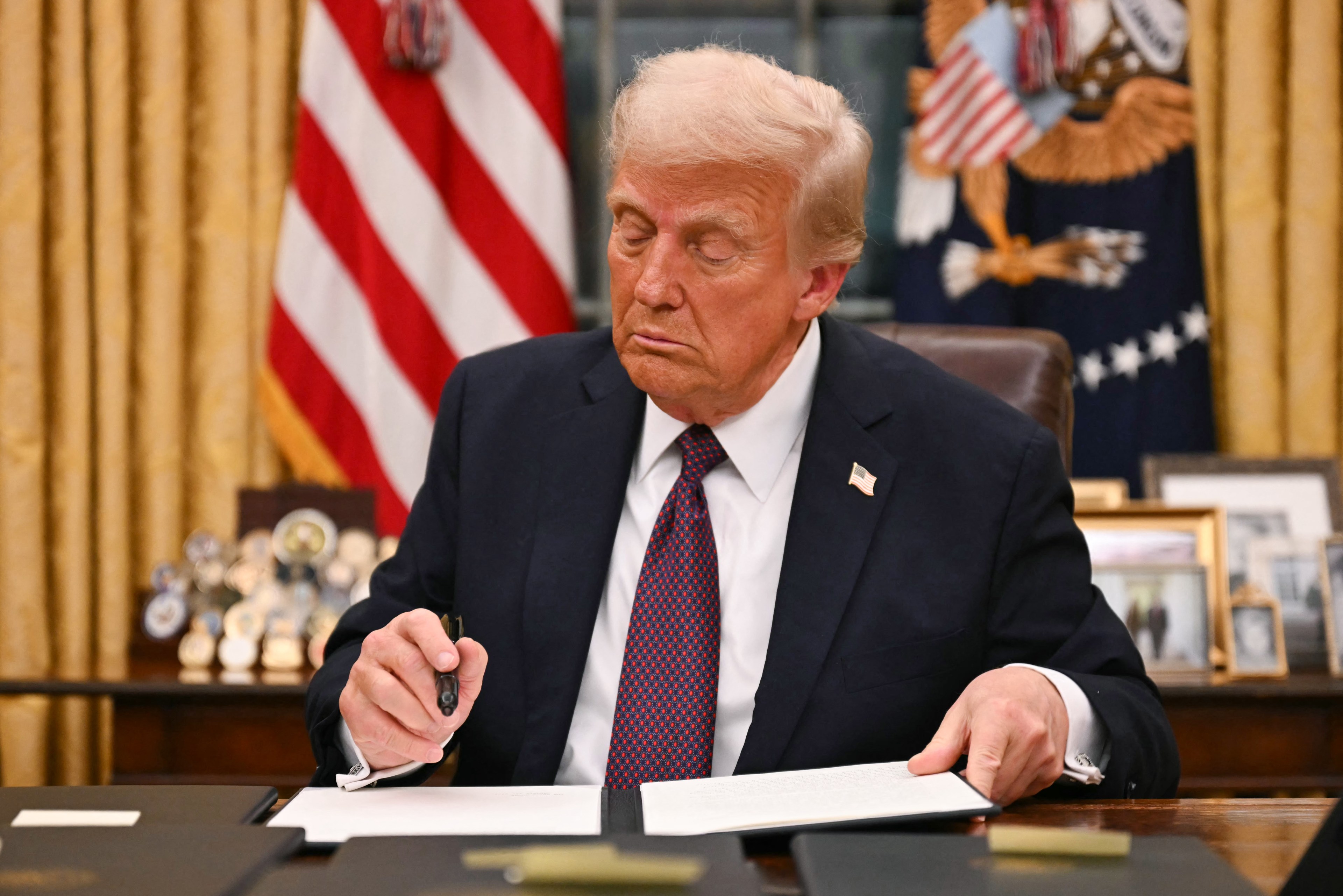 A man in a suit signs a document at a desk in an office, with a US flag, framed photos, and medals behind him.