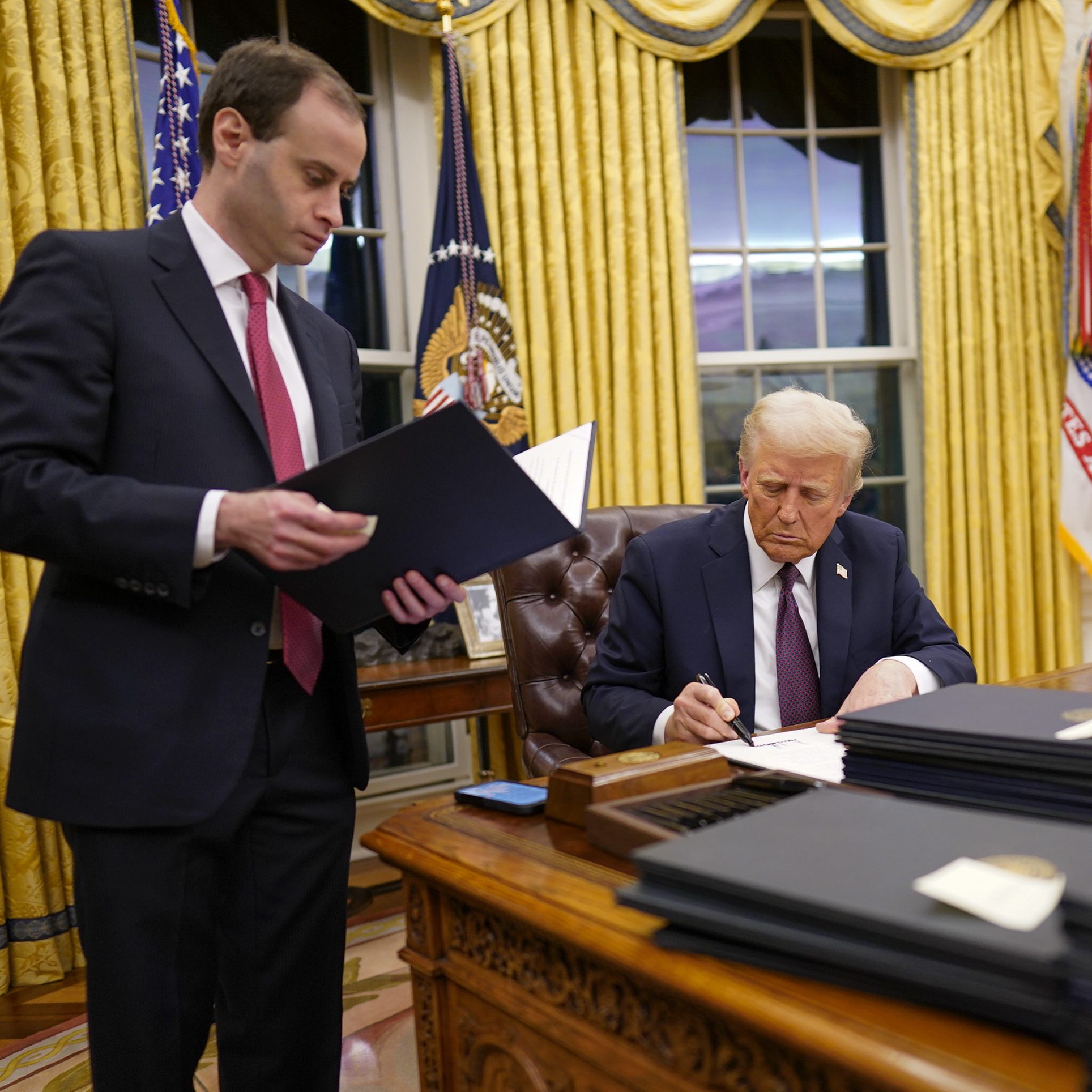 A man in a suit signs a document at a wooden desk, surrounded by stacks of folders. Another man stands nearby holding an open folder, in a room with yellow curtains.