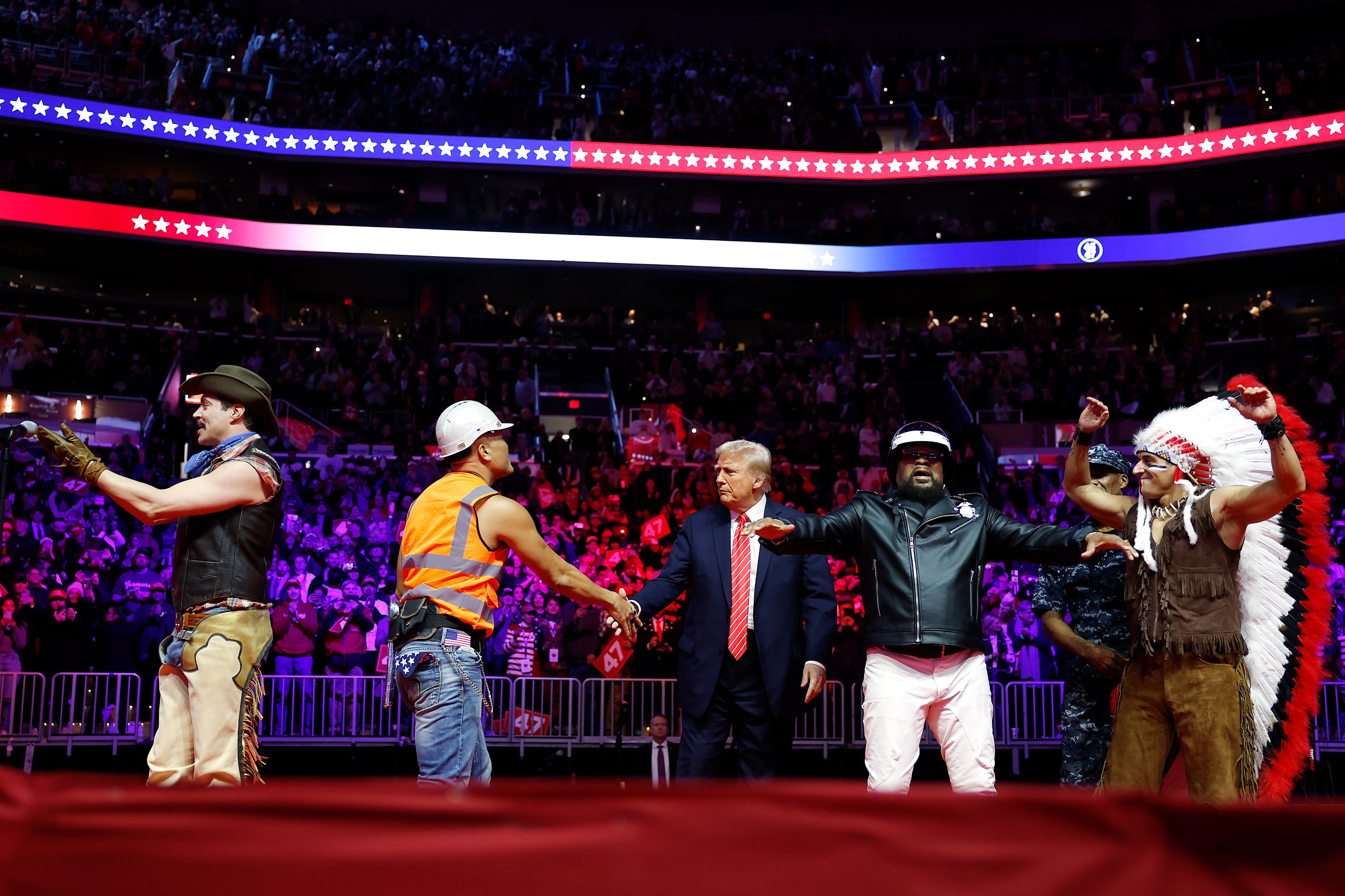 A group of performers in themed costumes stands in an arena. One person in a suit shakes hands with someone dressed as a construction worker. The audience watches.