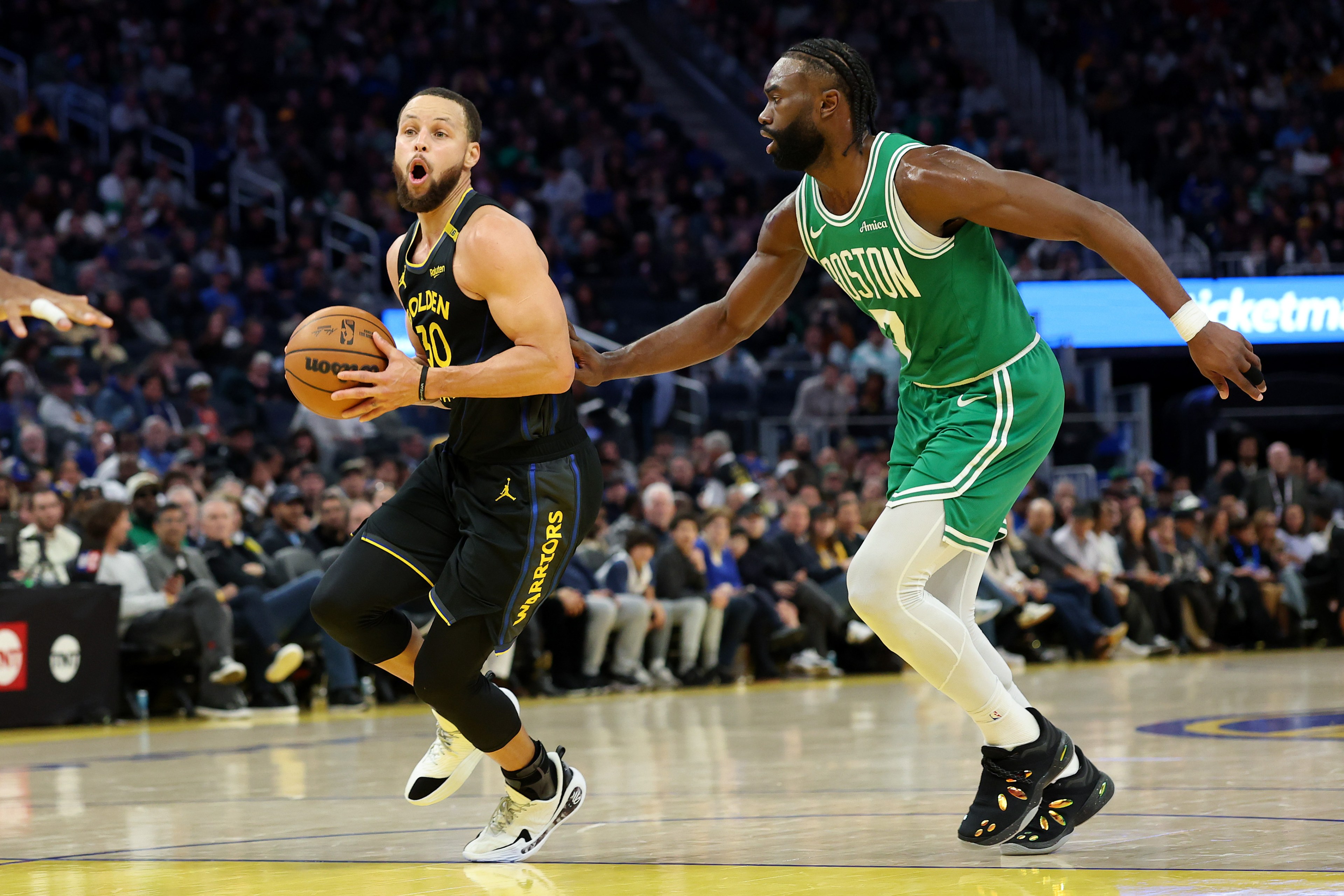 Two basketball players are in action on a court; one in a black "Golden" jersey dribbles, while the other in a green "Boston" jersey defends closely.