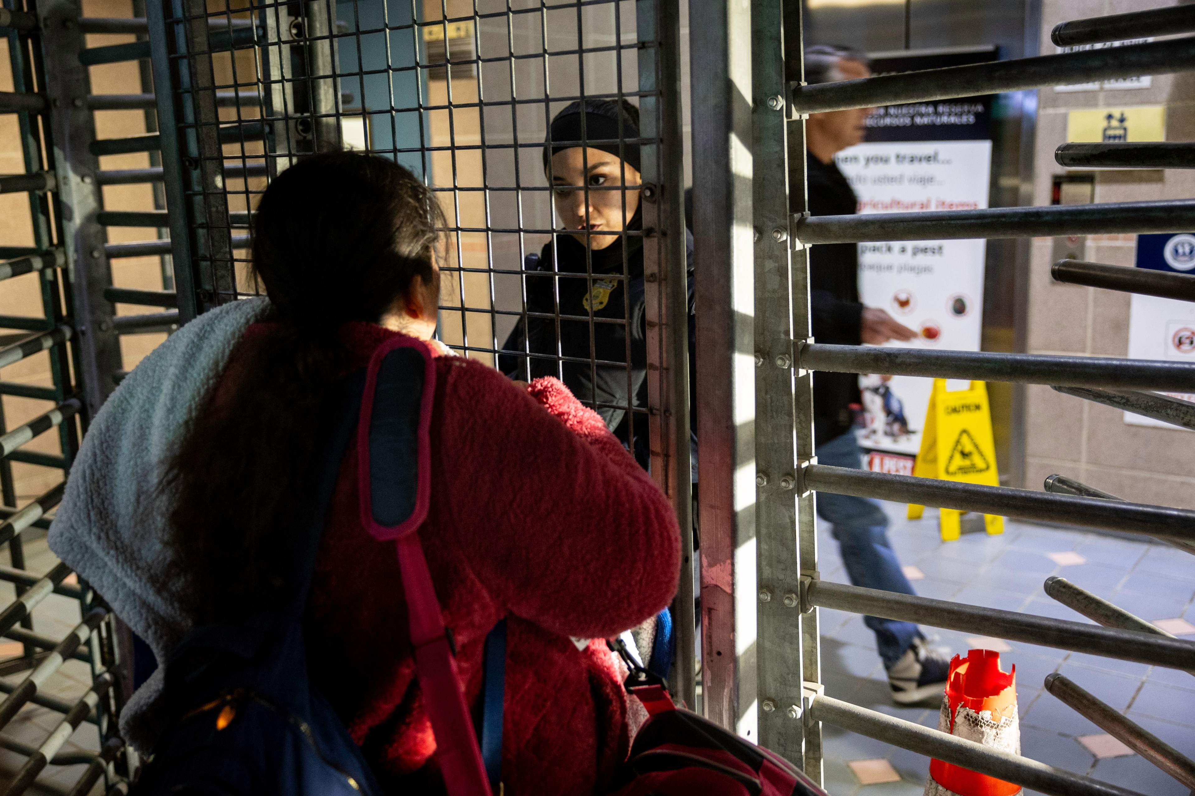 A woman in a red coat with bags faces a security officer behind a metal turnstile. The setting is indoors with caution signs and posters visible in the background.