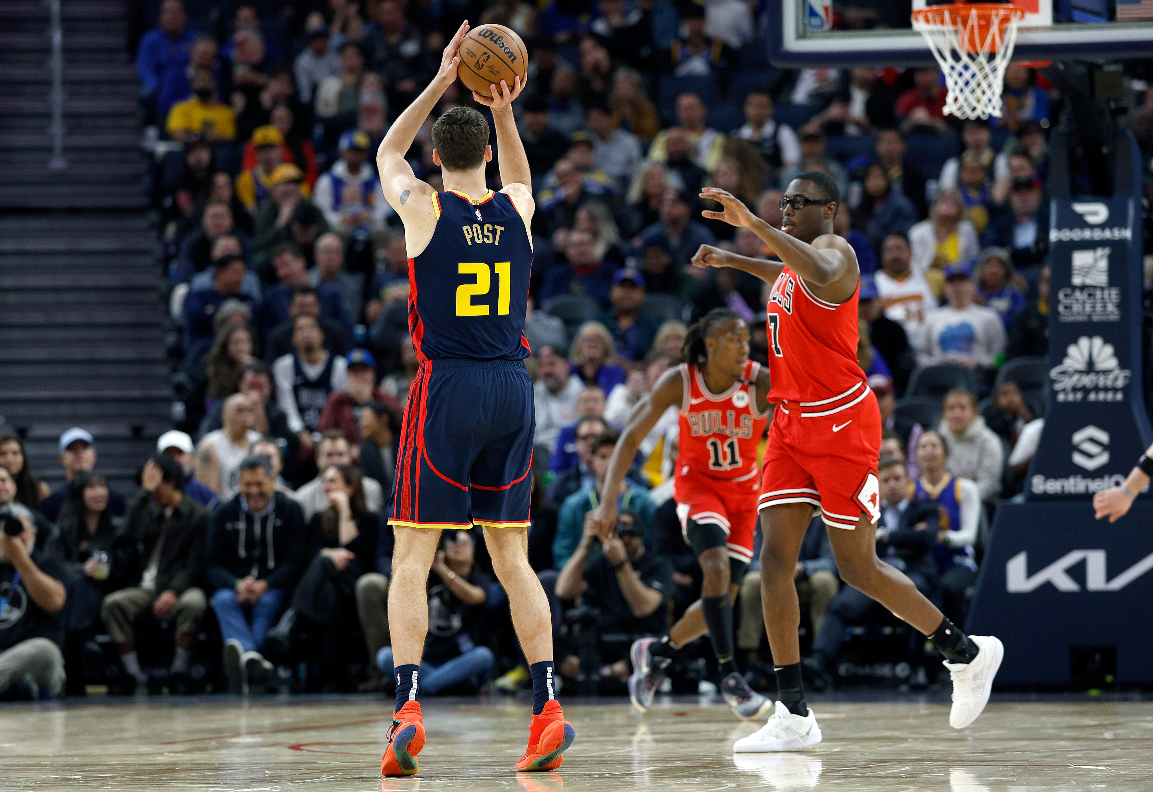 A basketball player in a navy jersey labeled &quot;21&quot; is shooting the ball while two opposing players in red jerseys attempt to block him. The crowd watches intently.