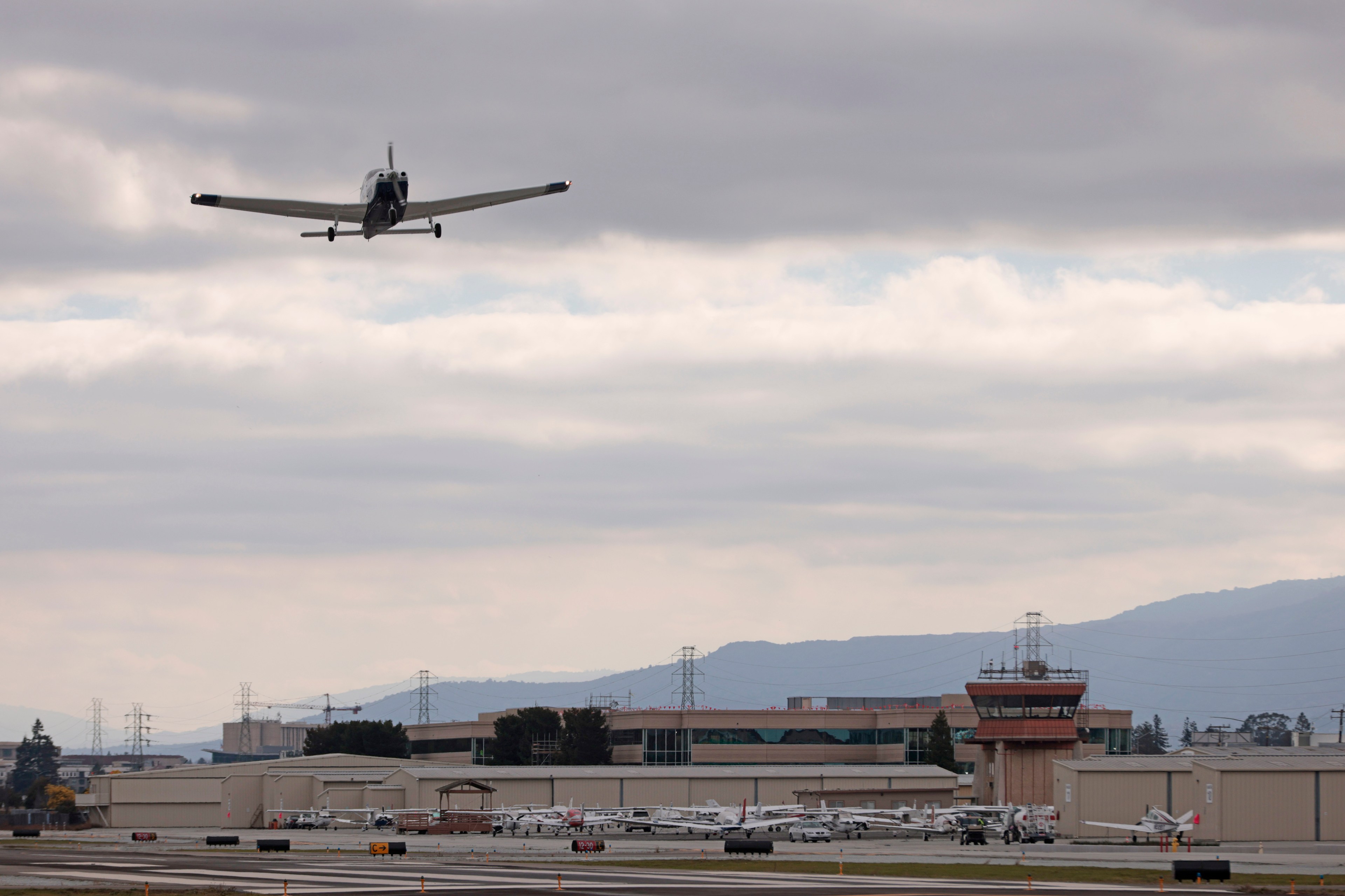 A small airplane flies above an airport runway with hangars and parked planes below, set against a cloudy sky and distant hills.