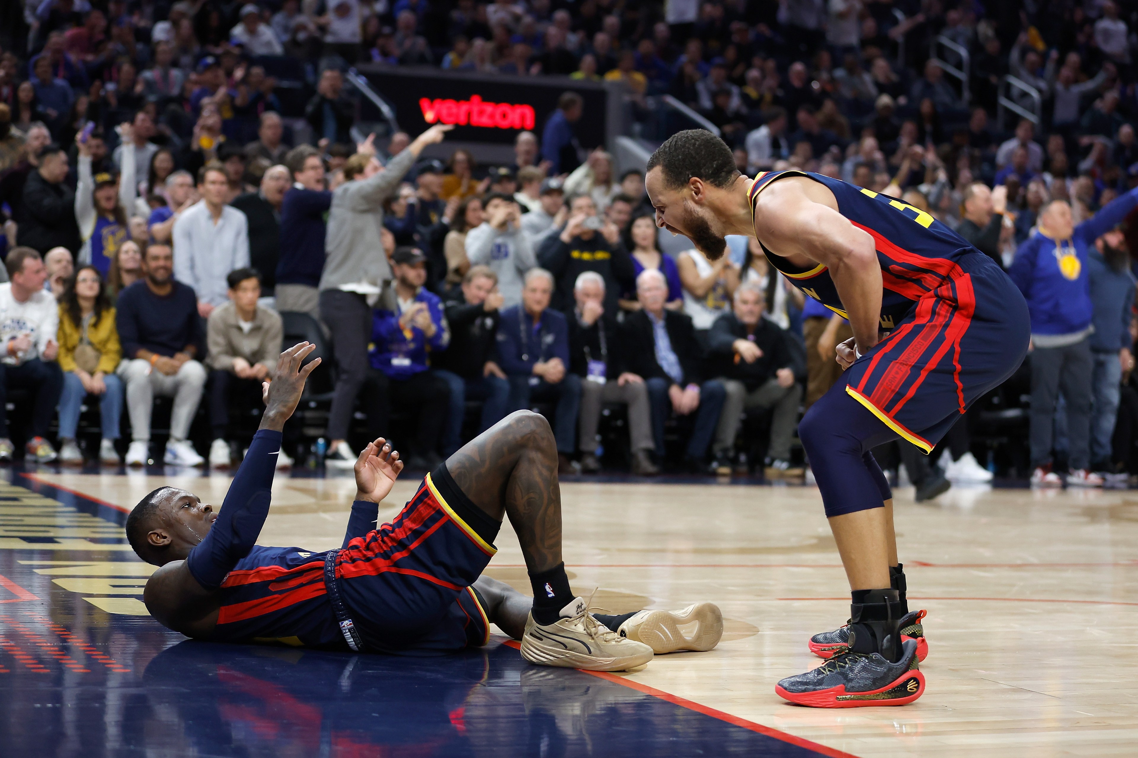 A basketball player is lying on the court, while another player in the same team's uniform is crouching and shouting energetically. A cheering crowd is in the background.