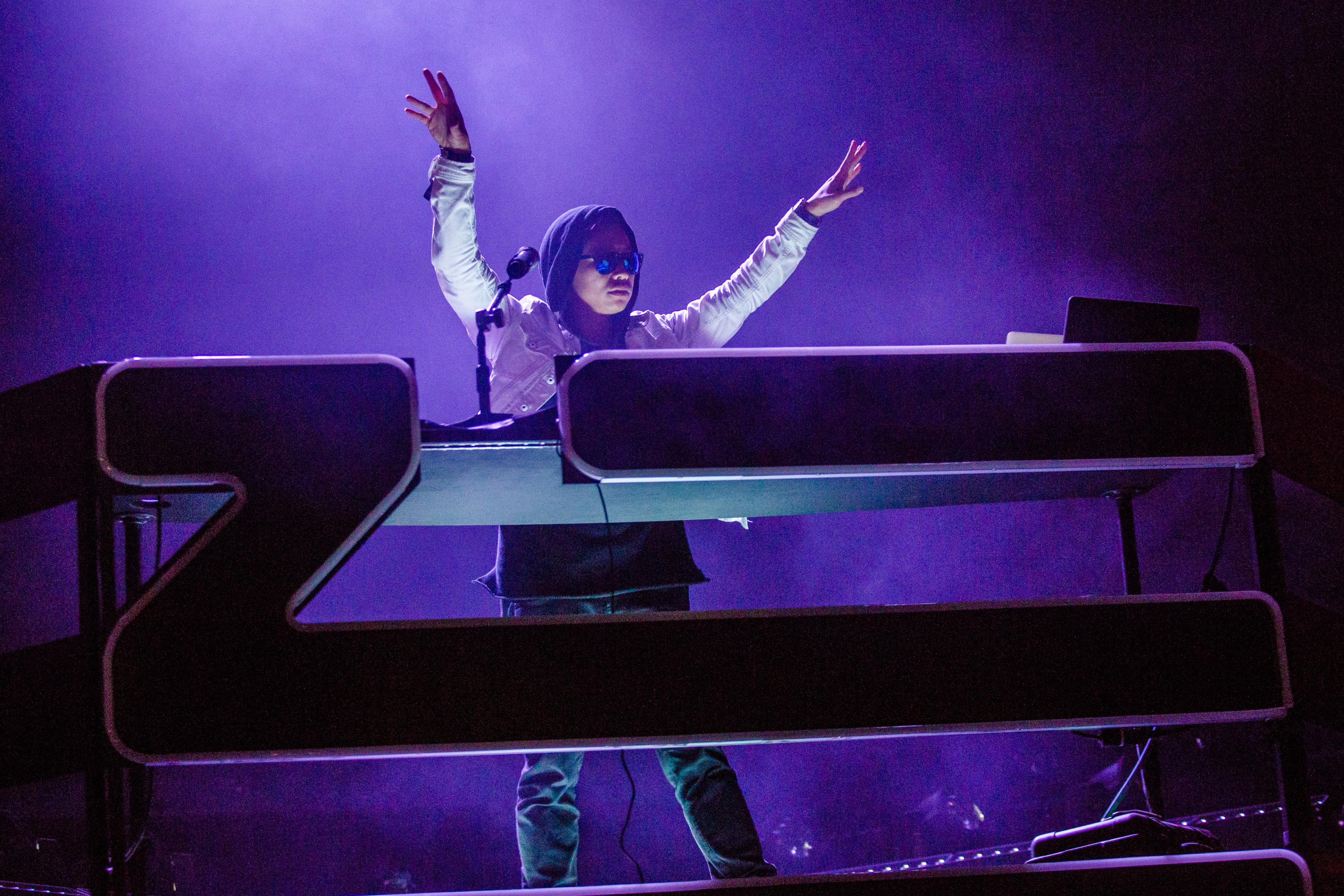 A DJ stands behind a large, illuminated console, wearing sunglasses and a hooded jacket with hands raised, against a backdrop of purple lights and mist.