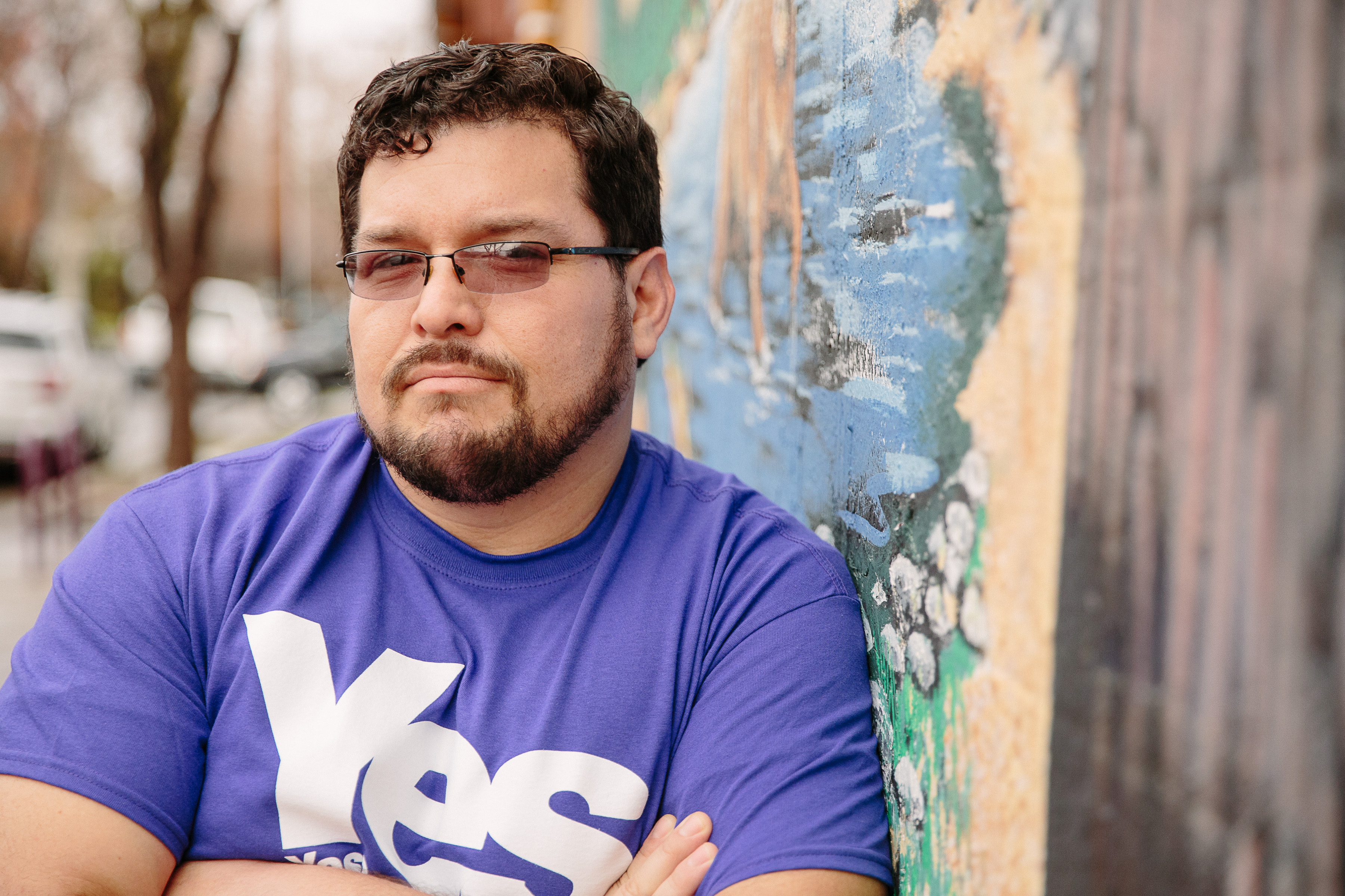 A person with glasses and a beard leans against a colorful mural. They are wearing a purple shirt with the word "Yes" prominently displayed.