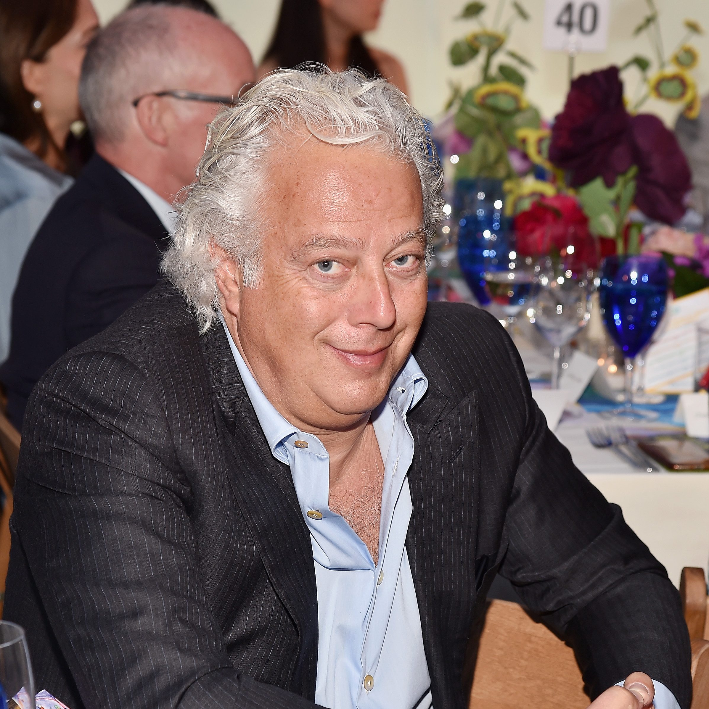 A smiling man with white hair and a light blue shirt under a black suit jacket is sitting at an elegantly set table with floral decorations and blue glasses.