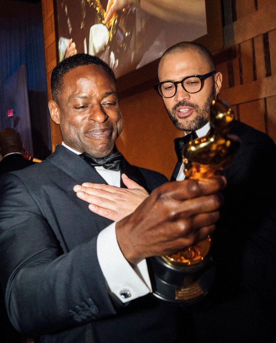 Two men in formal attire are happily admiring a golden statuette. One man holds it proudly while the other looks on with a warm smile.