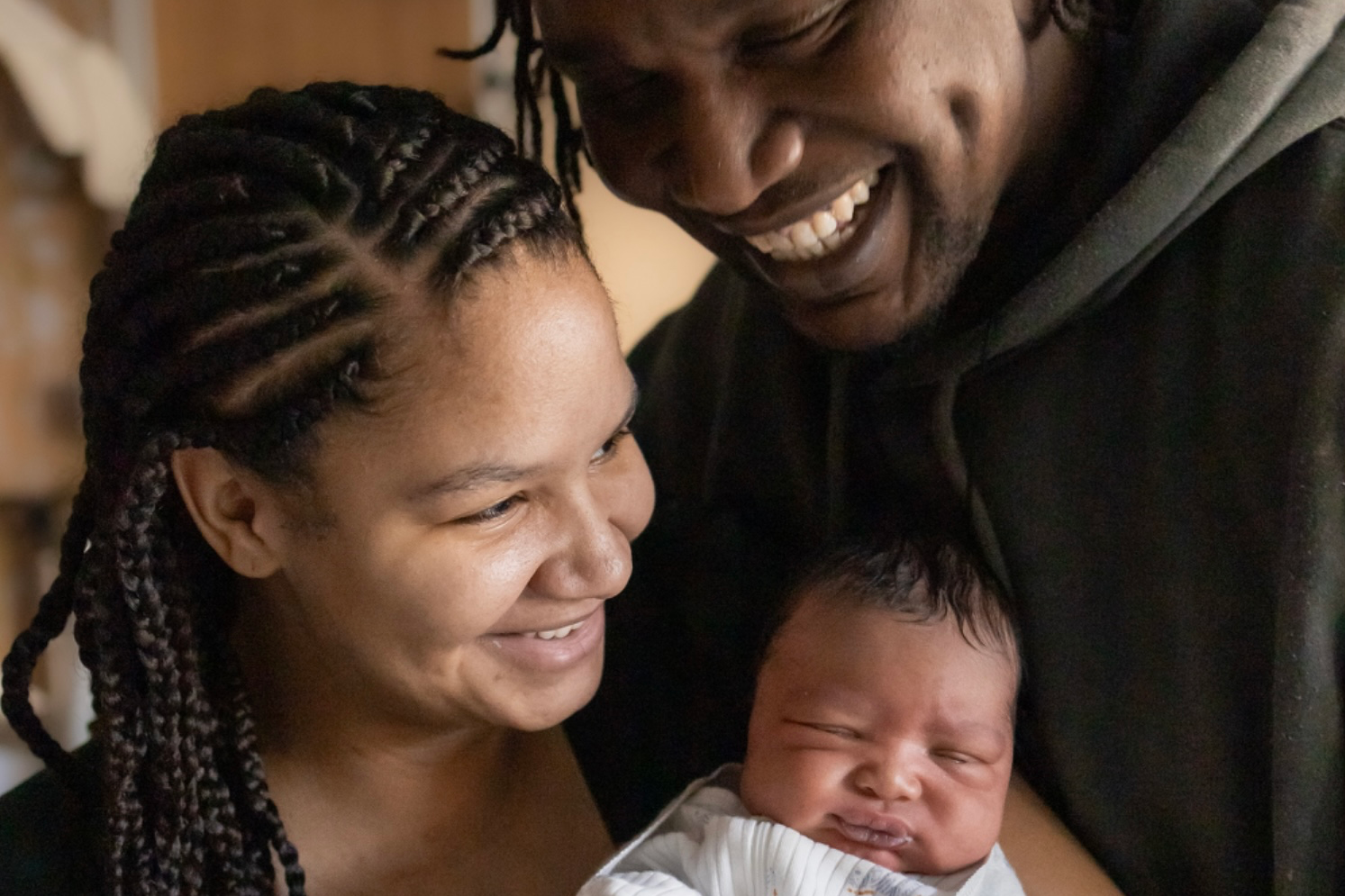 A joyful couple holds a newborn baby wrapped in a blanket. They are smiling warmly at the baby, sharing a tender moment in a softly lit room.