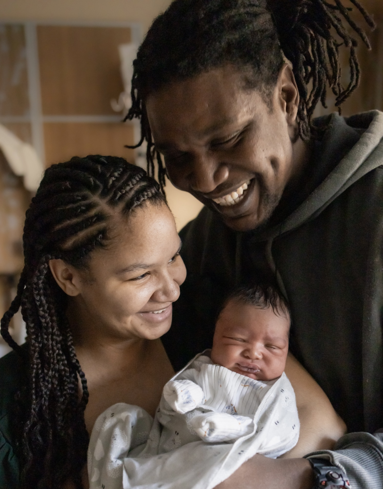 A joyful couple holds a newborn baby wrapped in a blanket. They are smiling warmly at the baby, sharing a tender moment in a softly lit room.