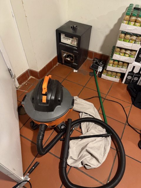 A shop vac with a hose lies on a tiled floor near a safe. Boxes of canned beverages are stacked nearby, along with a mop and a rolled-up cloth.