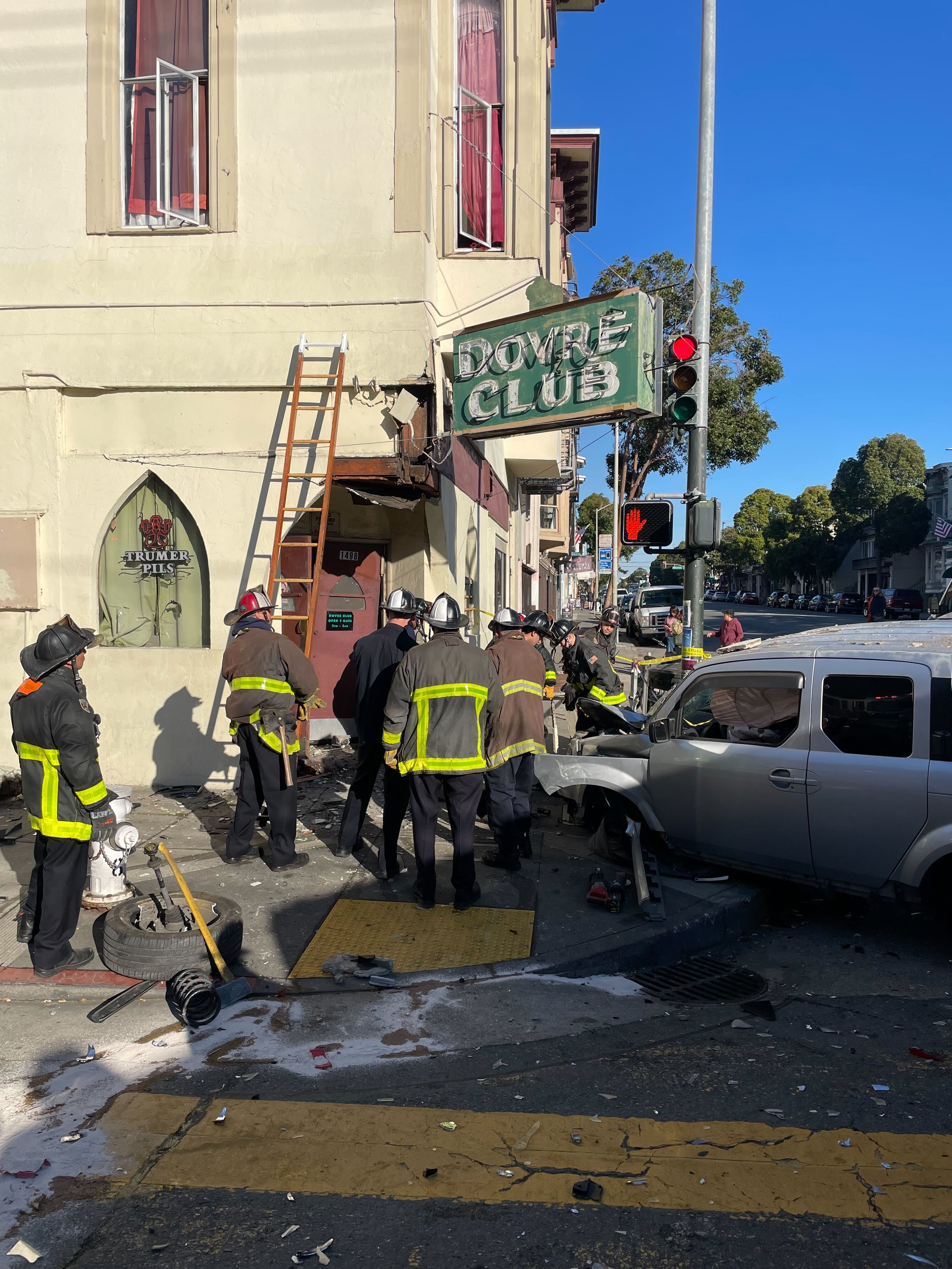 Firefighters and emergency responders surround a crashed SUV at the corner of a building marked "Dovre Club." Debris is scattered on the ground.