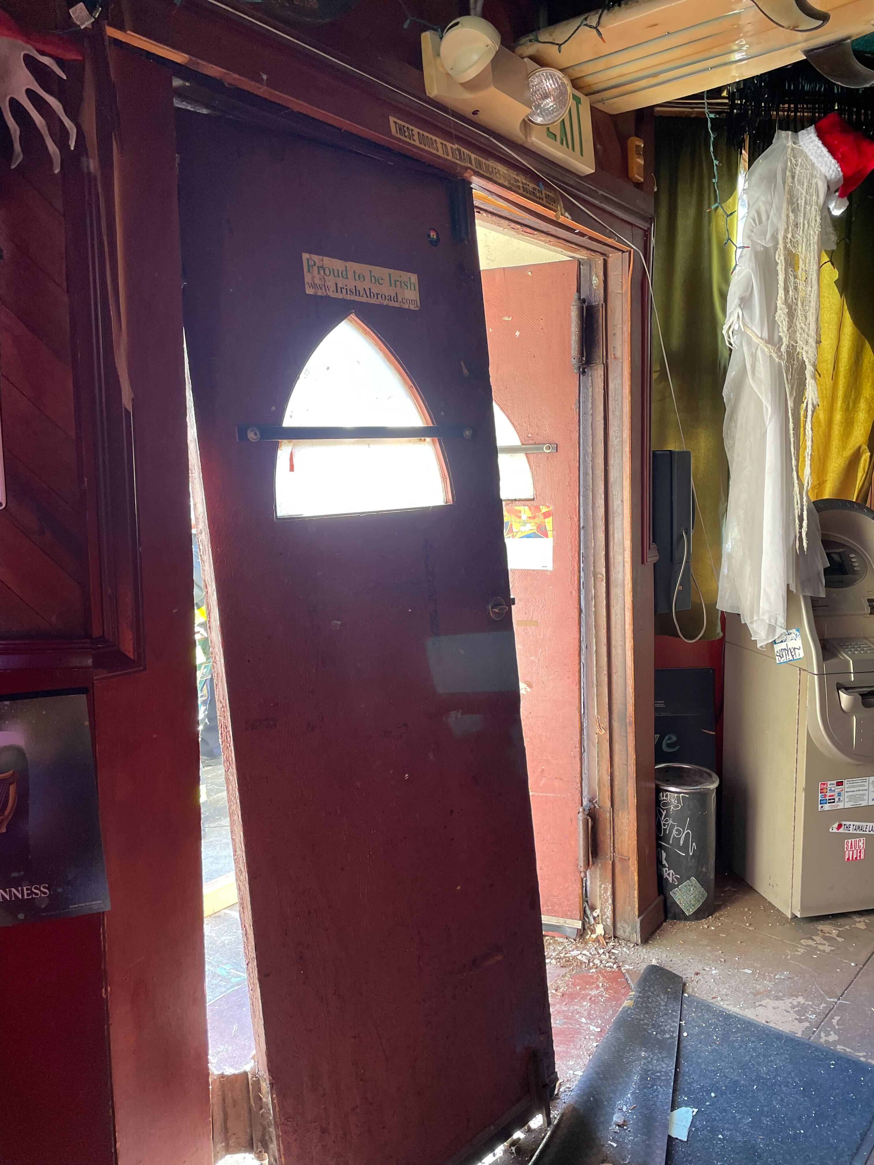 A wooden door with a sign reading &quot;Proud to be Irish&quot; is ajar, revealing sunlight. Nearby are an ATM, decorations, and a Santa hat on a draped figure.