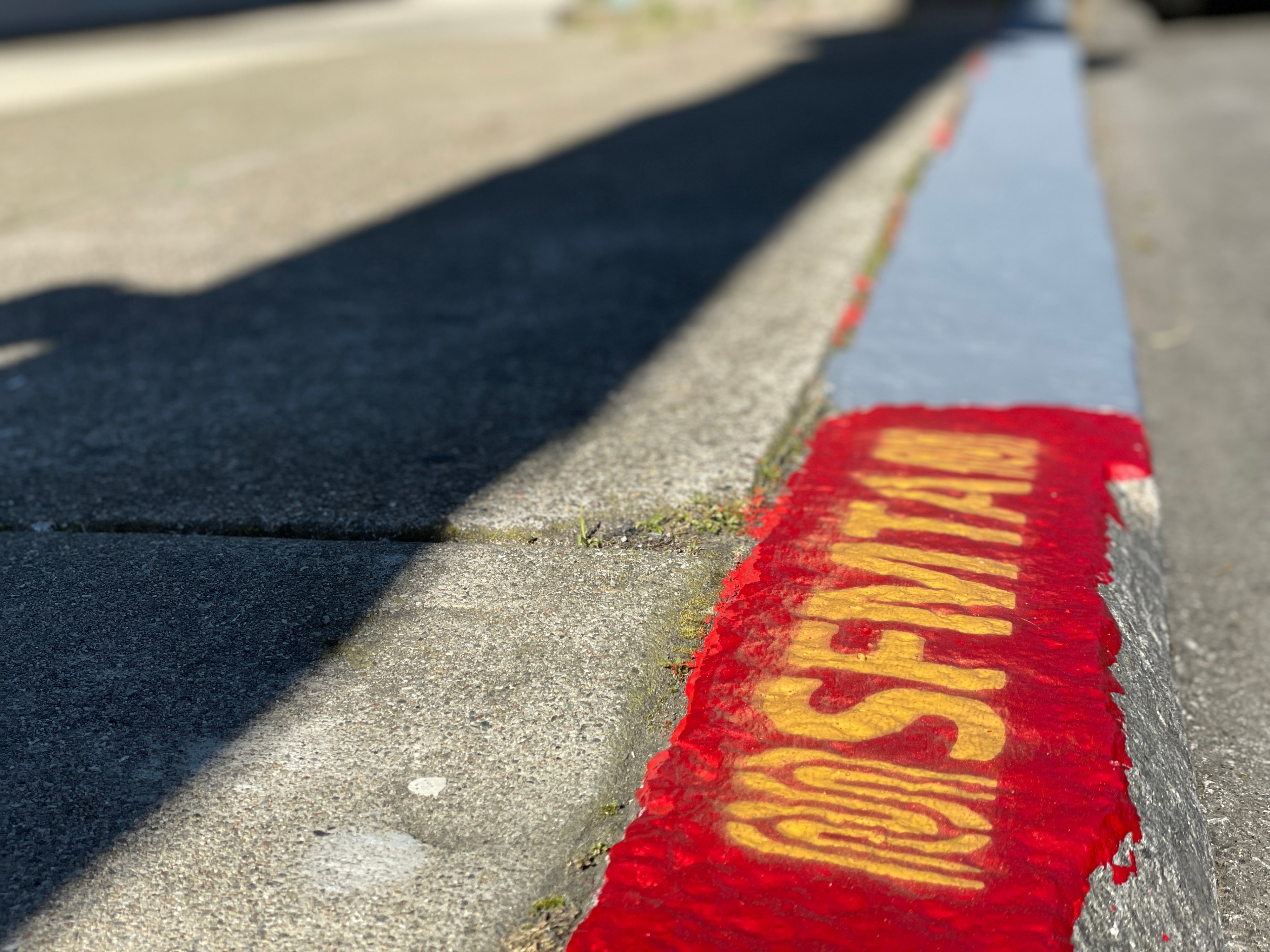 A sidewalk curb with "SFMTA" stenciled in yellow on red paint, casting shadows on the pavement in bright sunlight.