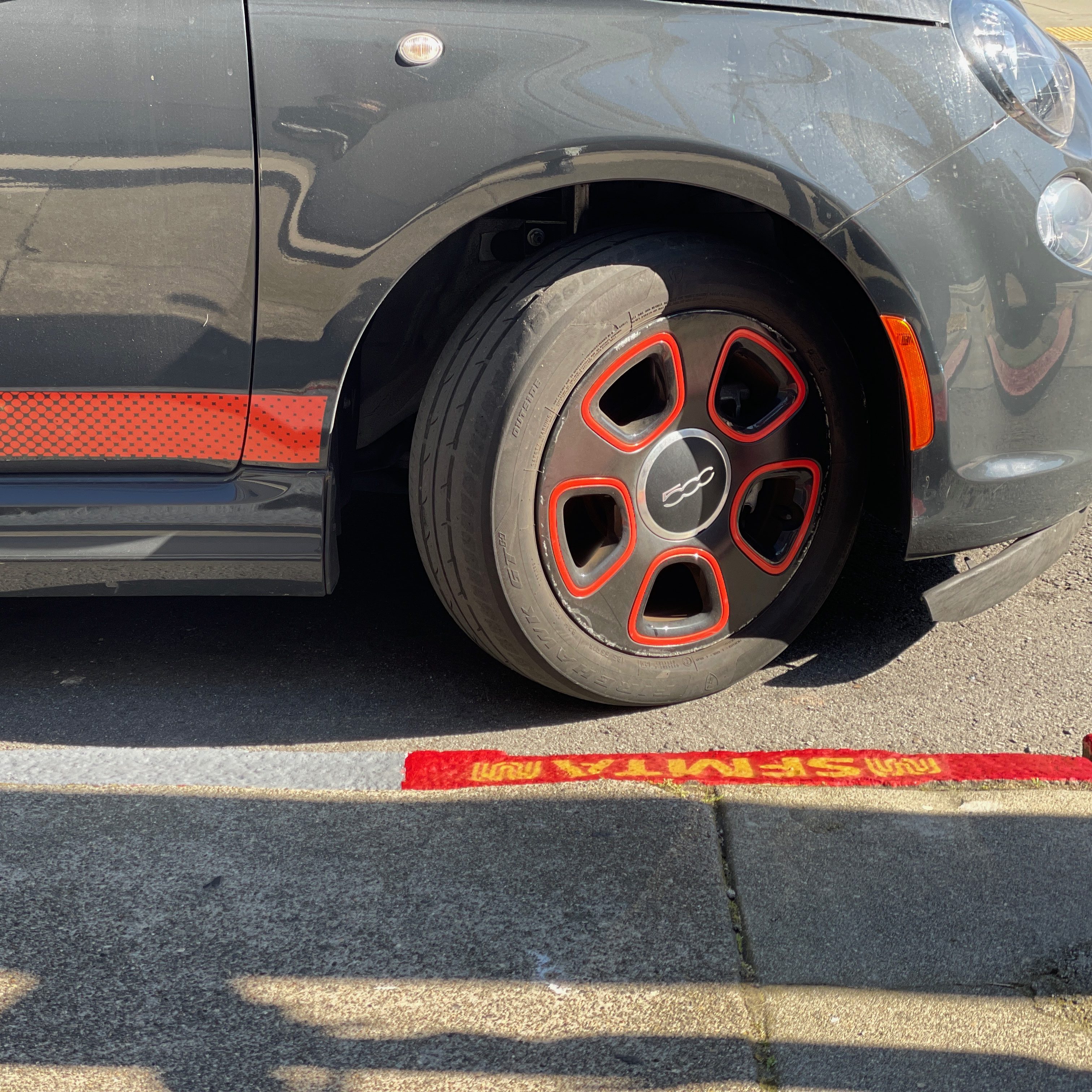 The image shows the front half of a small, dark gray car with red-trimmed wheel rims, partially parked on a street, next to a red curb marking.