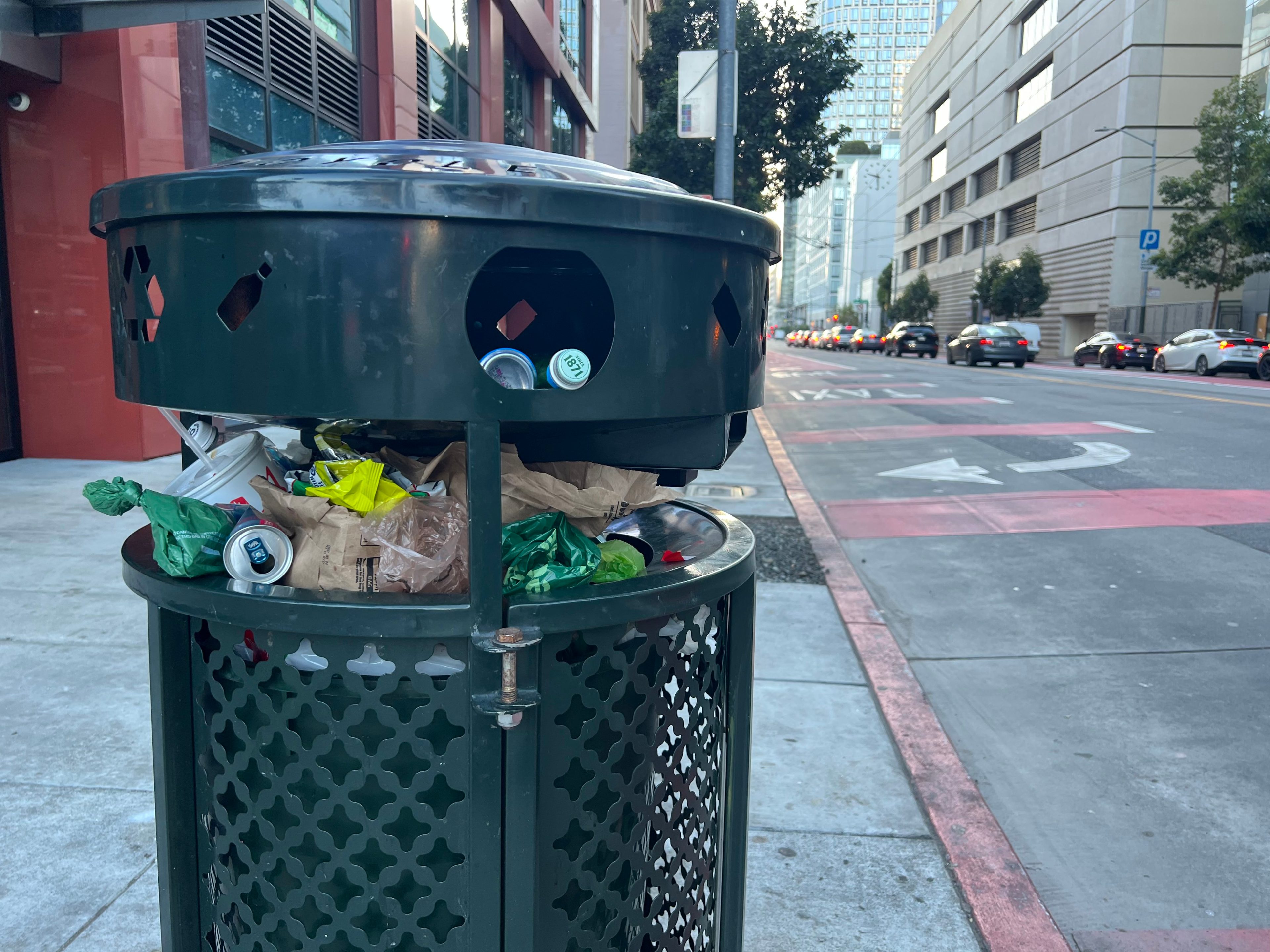 An overflowing trash can is on a city sidewalk, with buildings and a street full of cars in the background, under a clear sky.