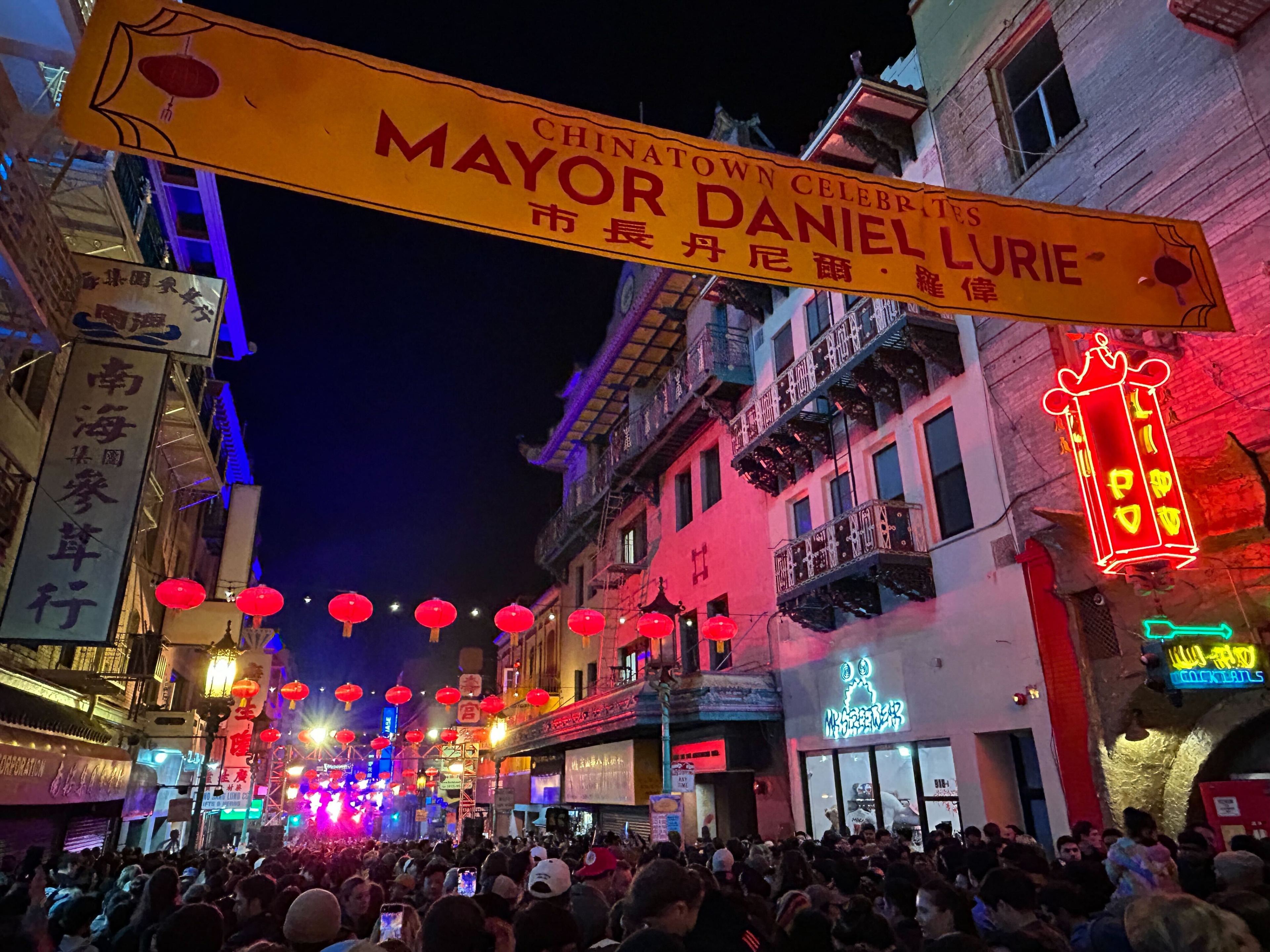 A crowded street in Chinatown is adorned with red lanterns and neon signs, with a banner celebrating Mayor Daniel Lurie above the bustling crowd.