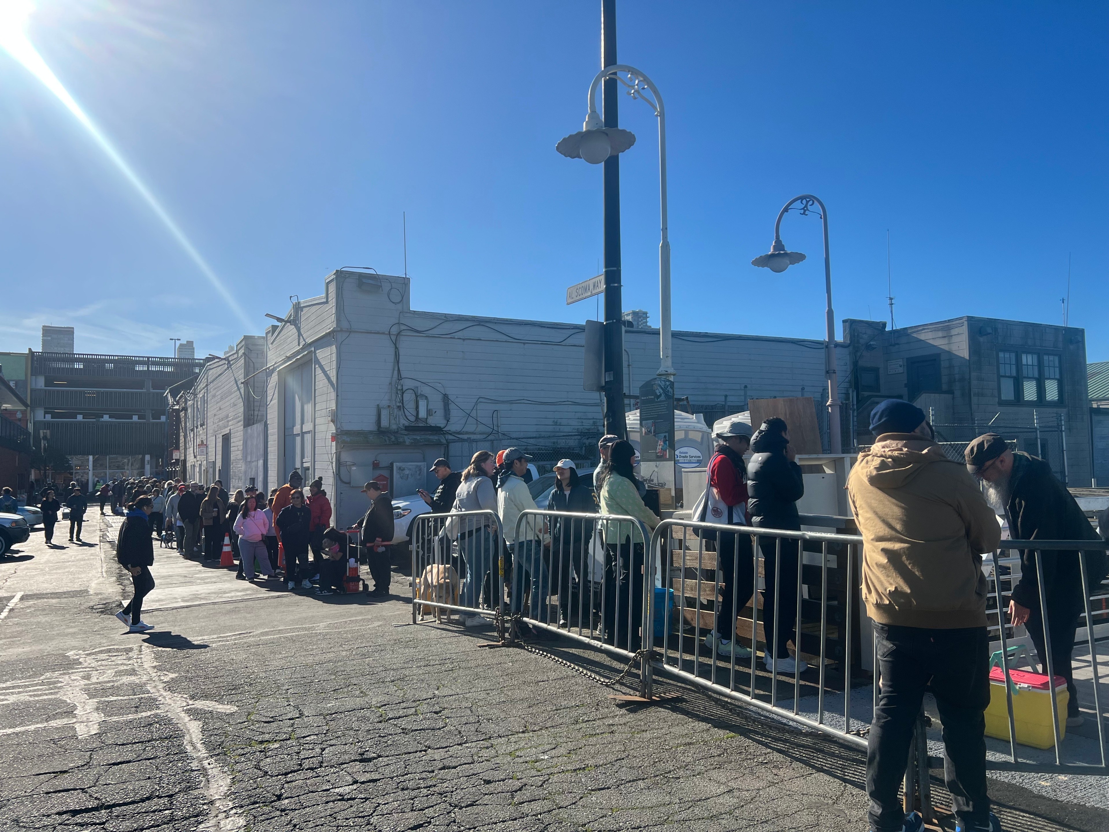 A long line of people stands outside near a building under a clear blue sky, bordered by metal barriers. Some individuals wear jackets and hats, suggesting cool weather.