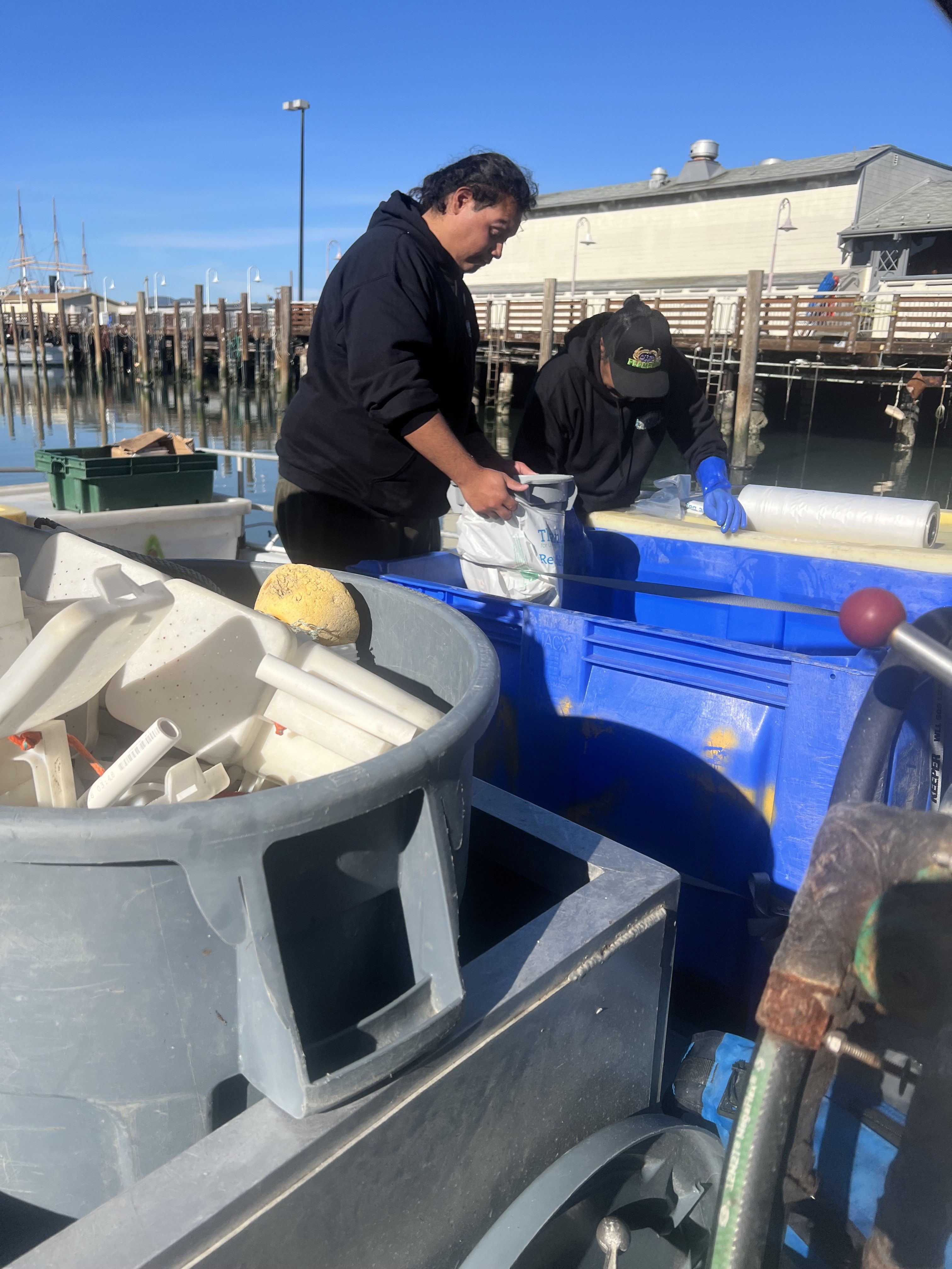 Two people in black hoodies work near large containers and bins filled with plastic items by a waterfront, under a clear blue sky.