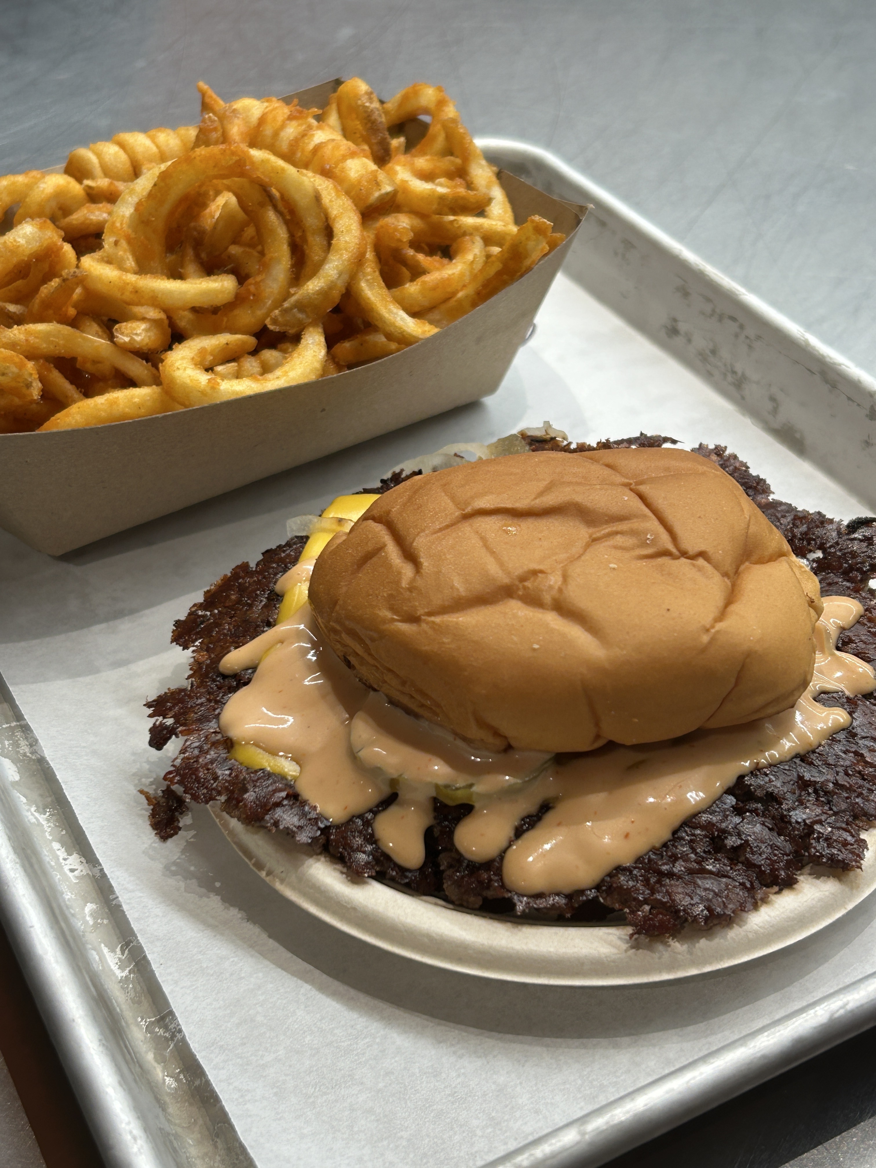 A hamburger with a toasted bun and sauce sits alongside a tray of seasoned curly fries on a metal tray lined with parchment paper.