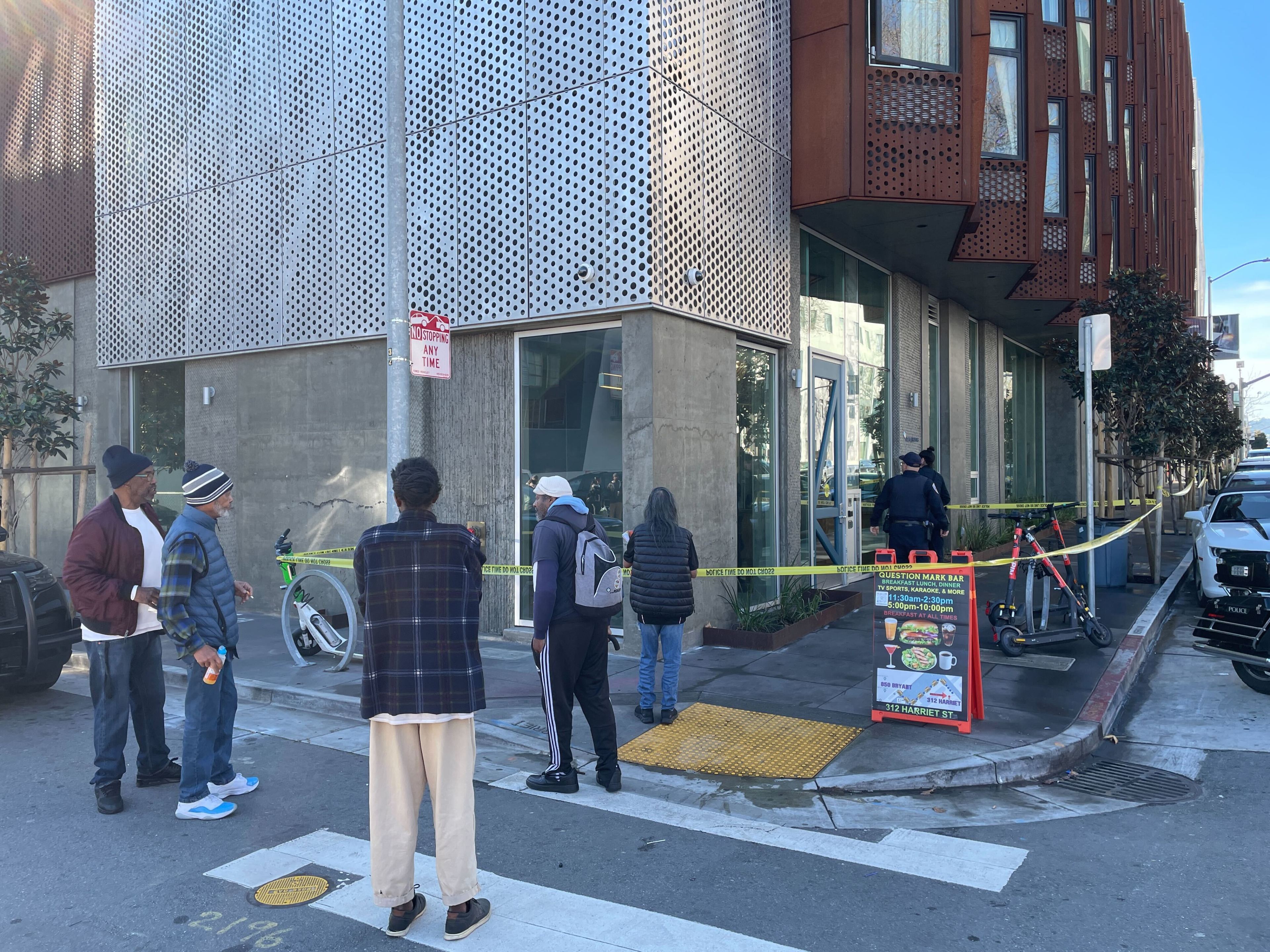 A group of people stands at a corner, observing a scene taped off by police. A few scooters and a bicycle are nearby, with a modern building in the background.