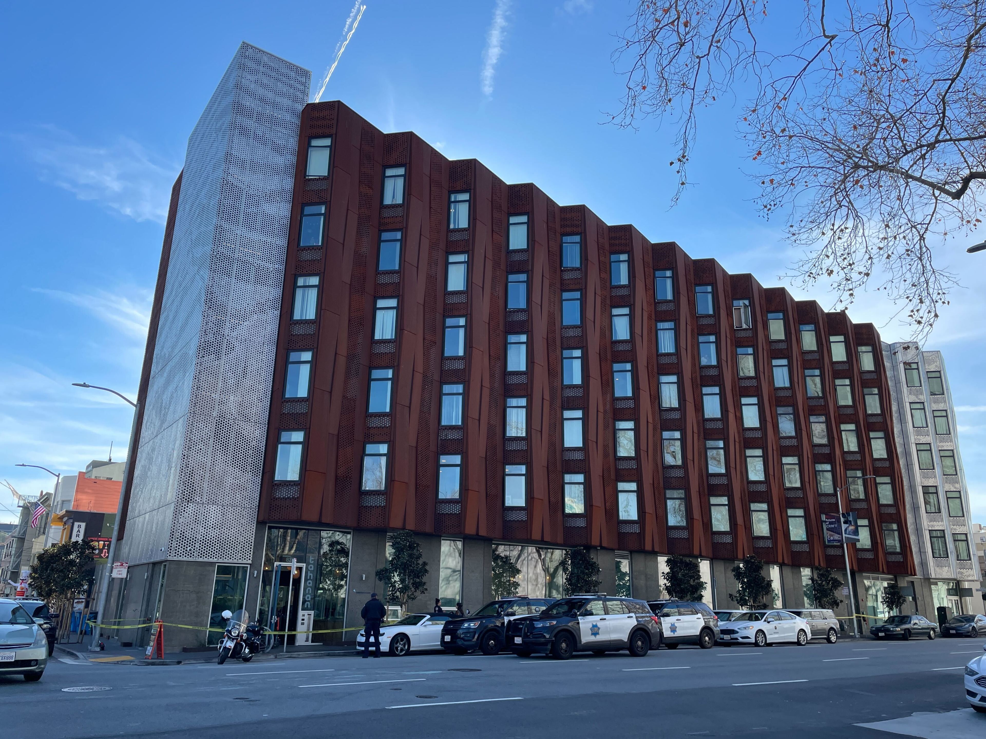 A modern, angular building with rusty-red facade towers next to a perforated metal structure. Police vehicles and a motorcycle are parked on the street nearby.