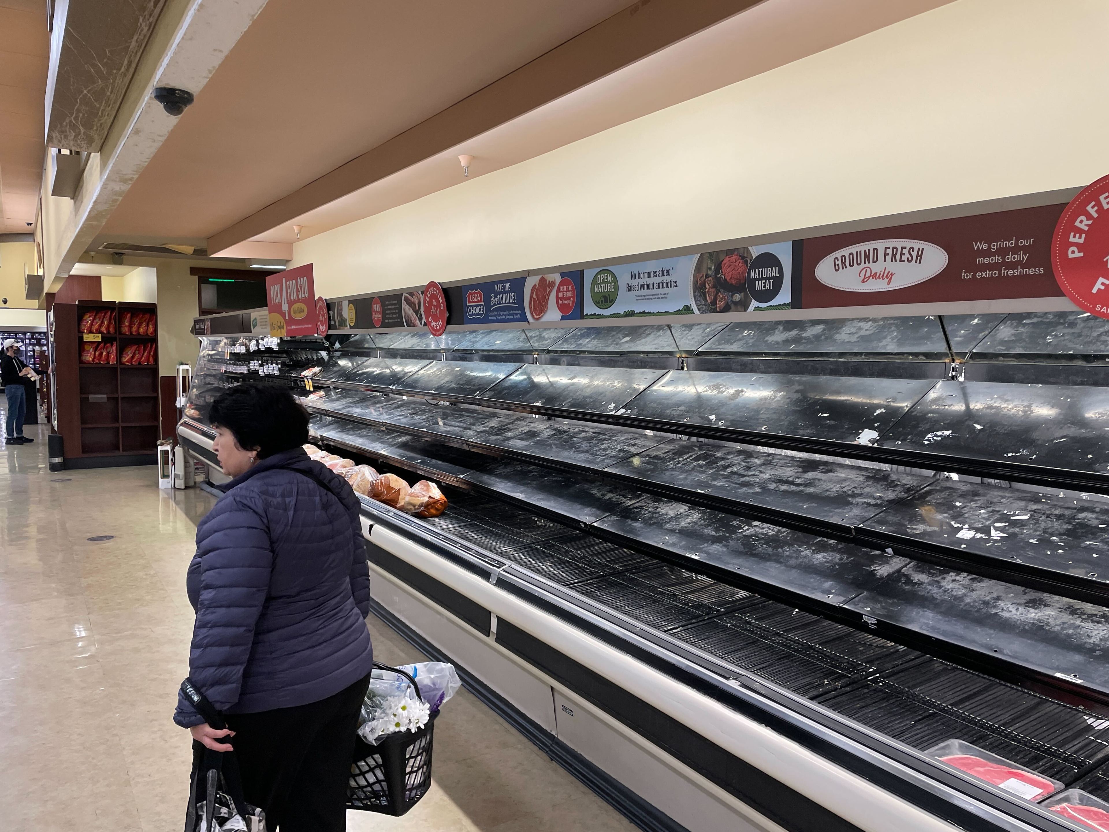 A person with a shopping basket stands near mostly empty supermarket shelves, with a few bread loaves displayed. Only a few items are visible in the aisle.