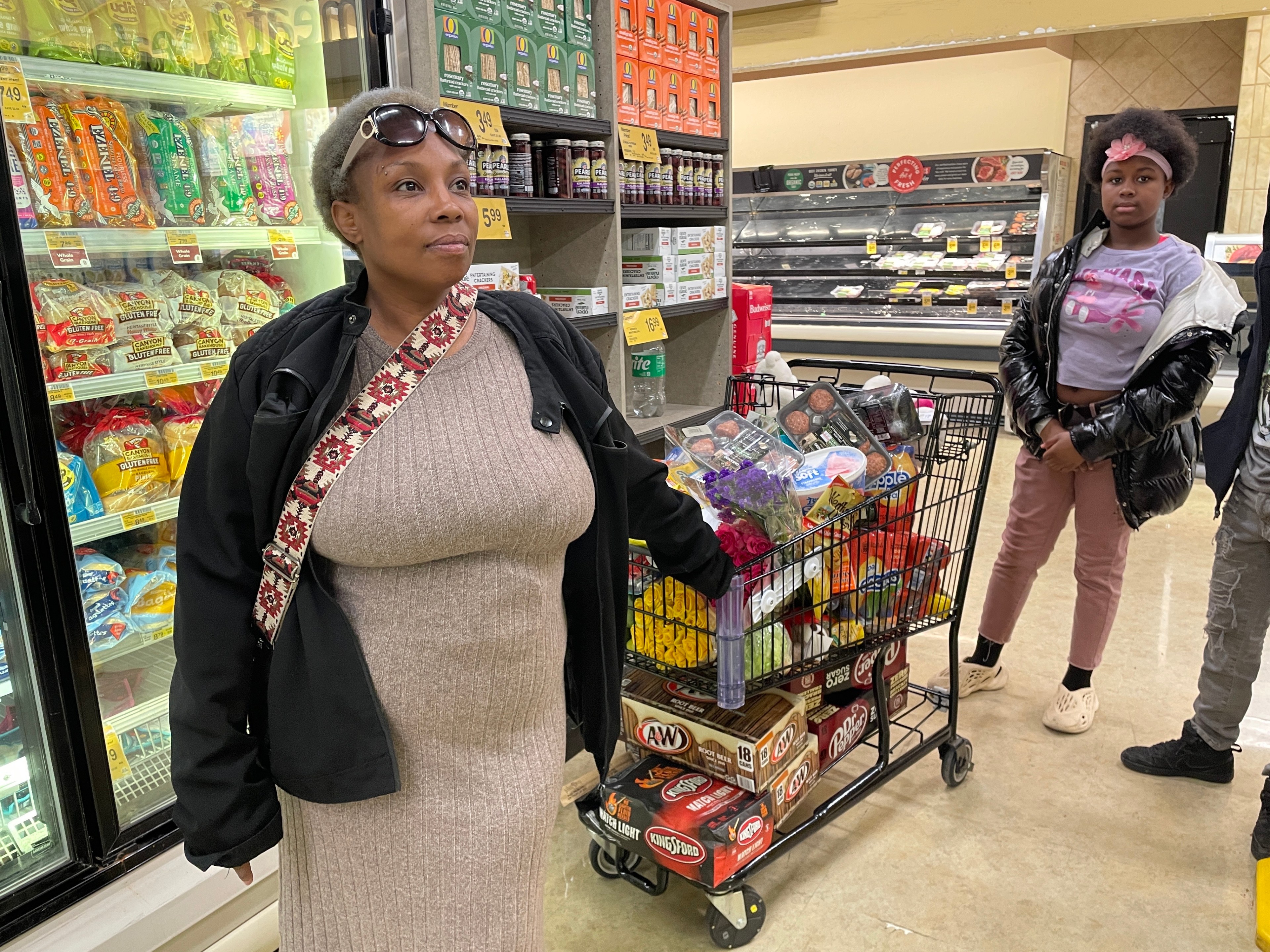 A woman stands near a grocery cart filled with items like soda, cookies, and flowers. Another person is nearby, with store shelves in the background.